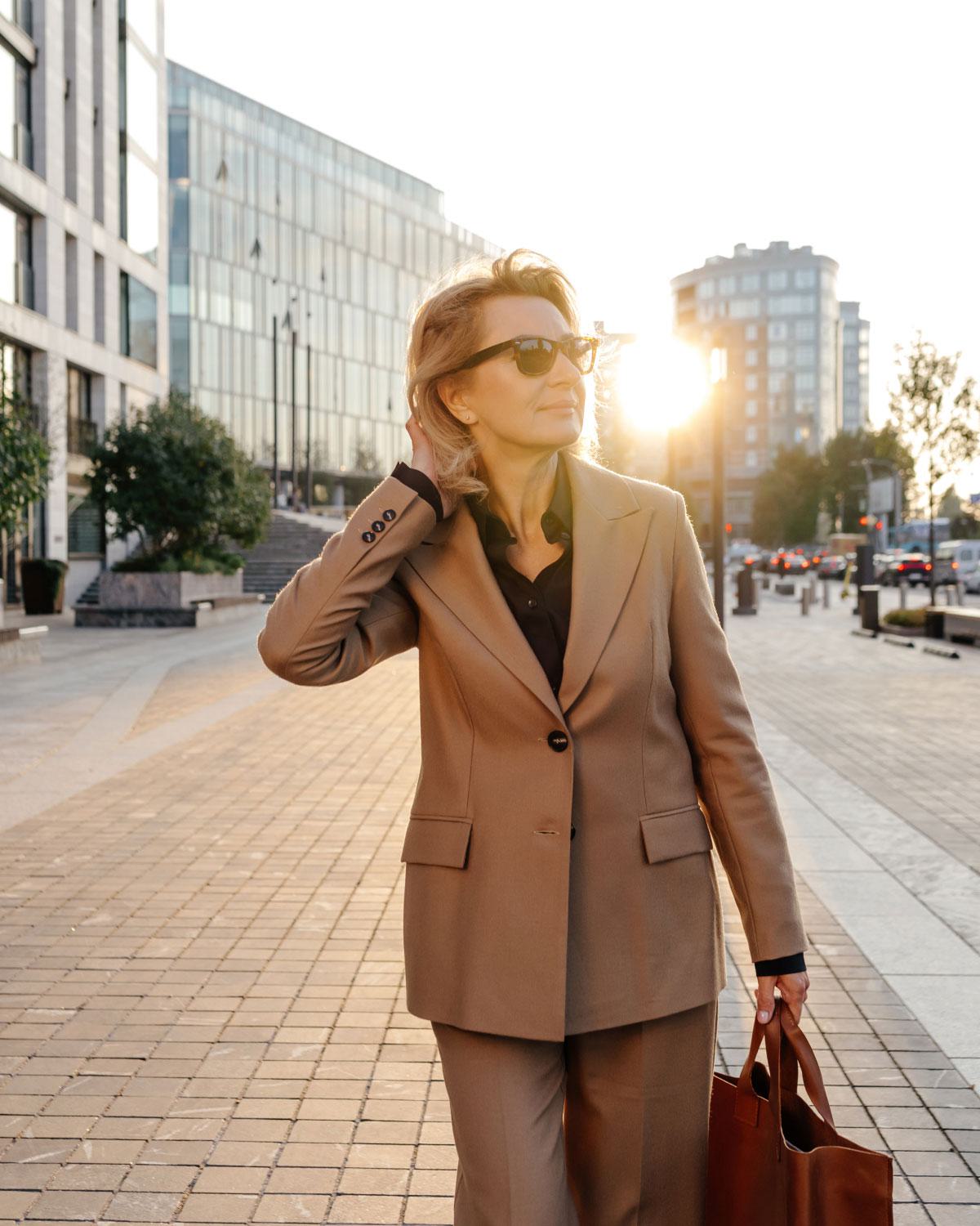 Confident woman in a tan suit and sunglasses walking through a modern urban plaza at sunset, carrying a leather tote bag