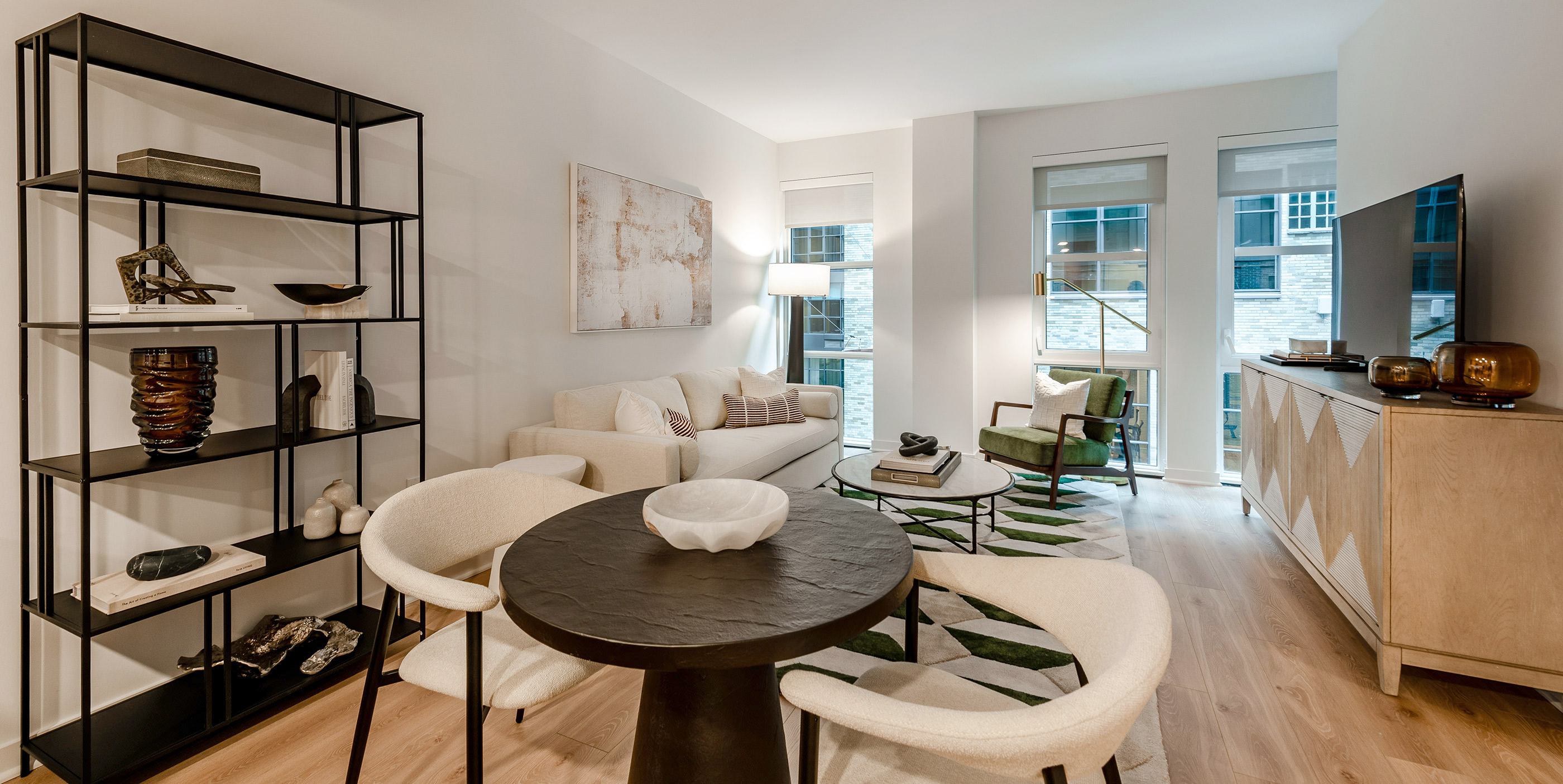 Modern living and dining area in a Nouvelle apartment, featuring minimalist furnishings, light wood floors, floor-to-ceiling windows, and curated shelving with contemporary décor accents.