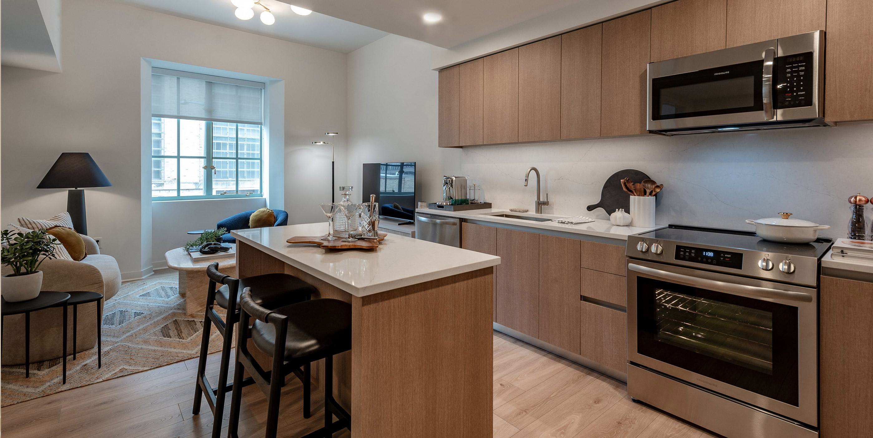 Modern kitchen and living area in a renovated apartment at Annex on 12th, featuring warm wood cabinetry, stainless steel appliances, a central island with bar seating, and a cozy lounge space near a large window.