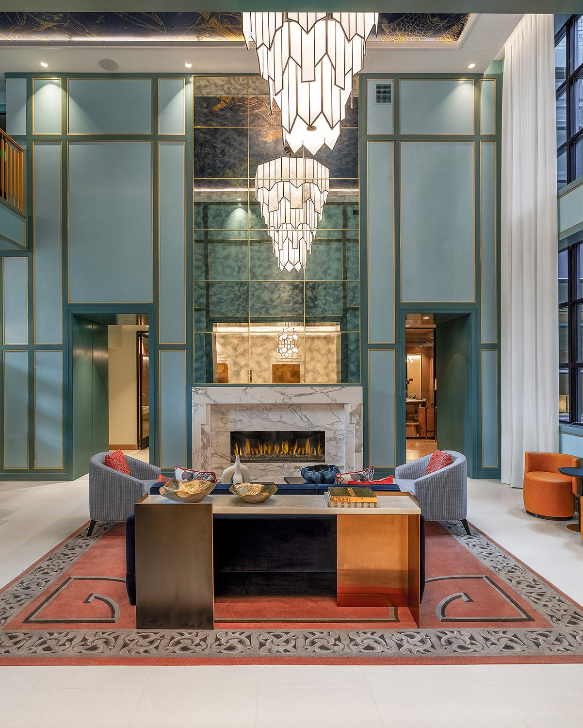 Grand lobby with soaring ceilings, teal and gold paneled walls, an elegant marble fireplace, and modern Art Deco chandeliers, featuring a mix of contemporary seating areas and tall windows that bring in natural light.