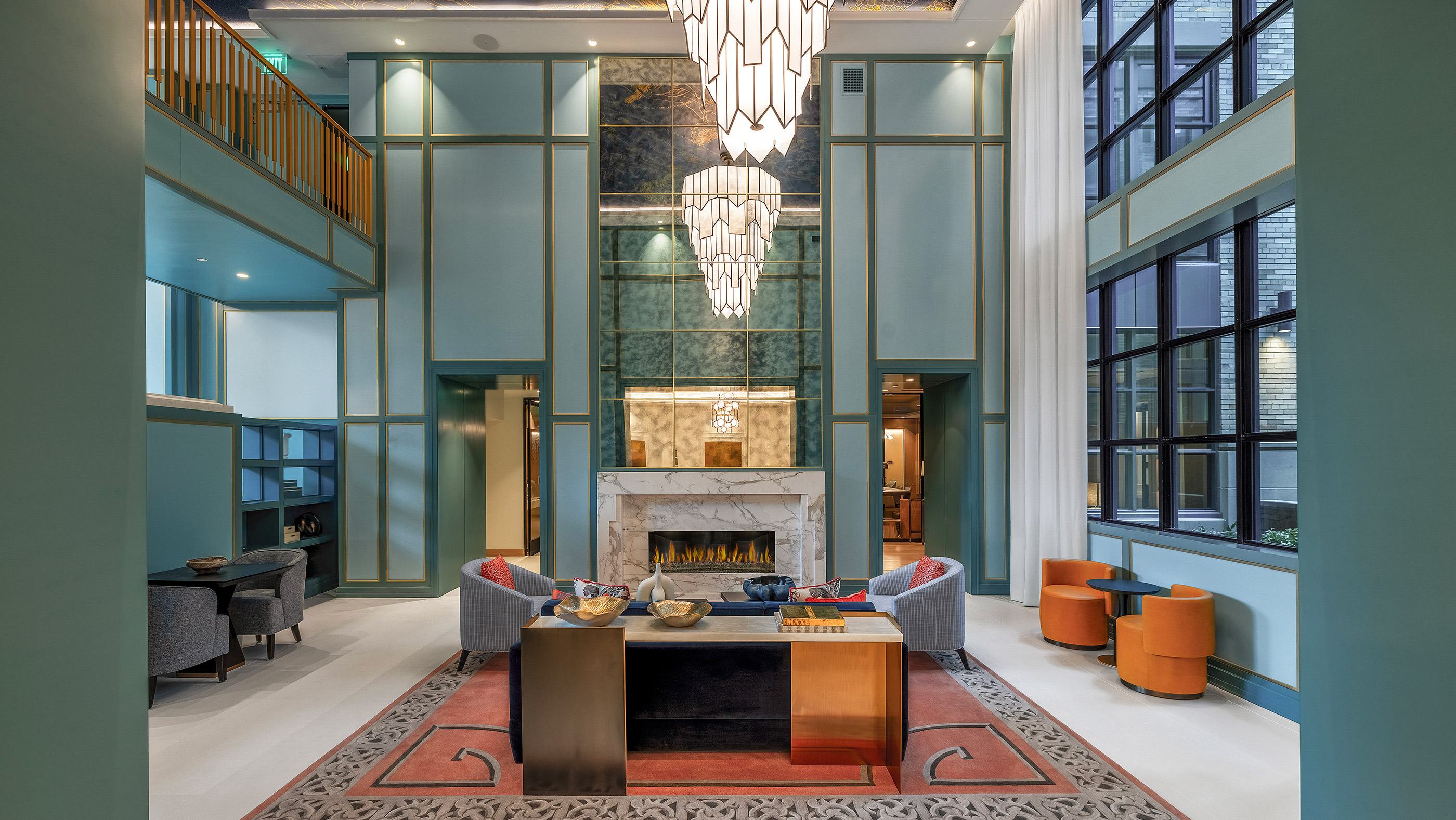 Grand lobby with soaring ceilings, teal and gold paneled walls, an elegant marble fireplace, and modern Art Deco chandeliers, featuring a mix of contemporary seating areas and tall windows that bring in natural light.