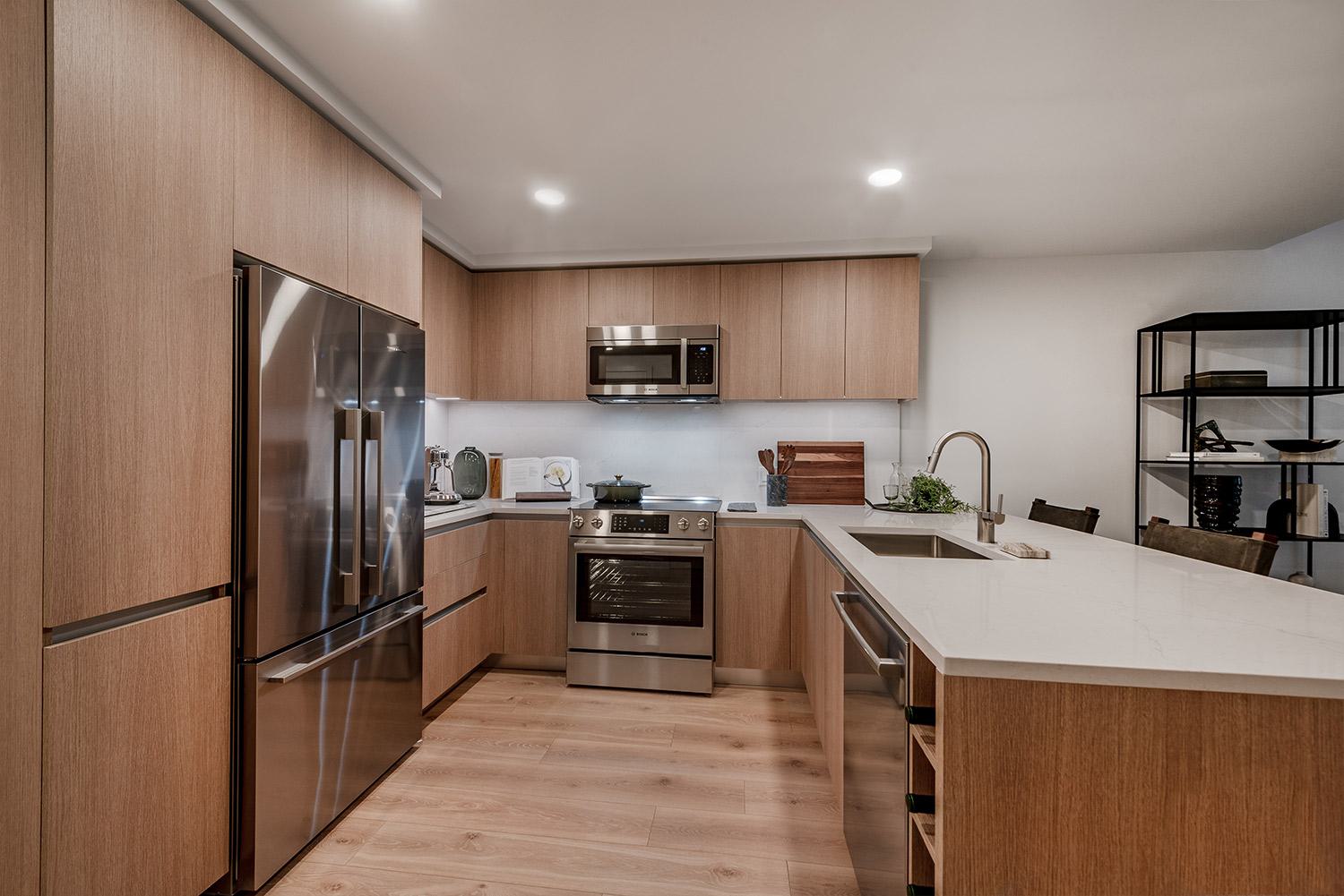 Modern kitchen at the Nouvelle building in the Annex on 12th, featuring warm wood cabinetry, stainless steel appliances, quartz countertops, and an oversized island with integrated sink and seating