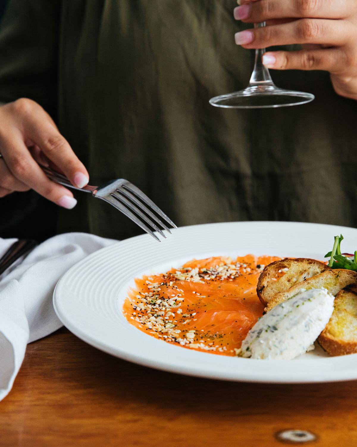 Close-up of a plate with cured salmon topped with seasoning, toast, a quenelle of herbed cream cheese, and a small salad garnish, with a person holding a wine glass.