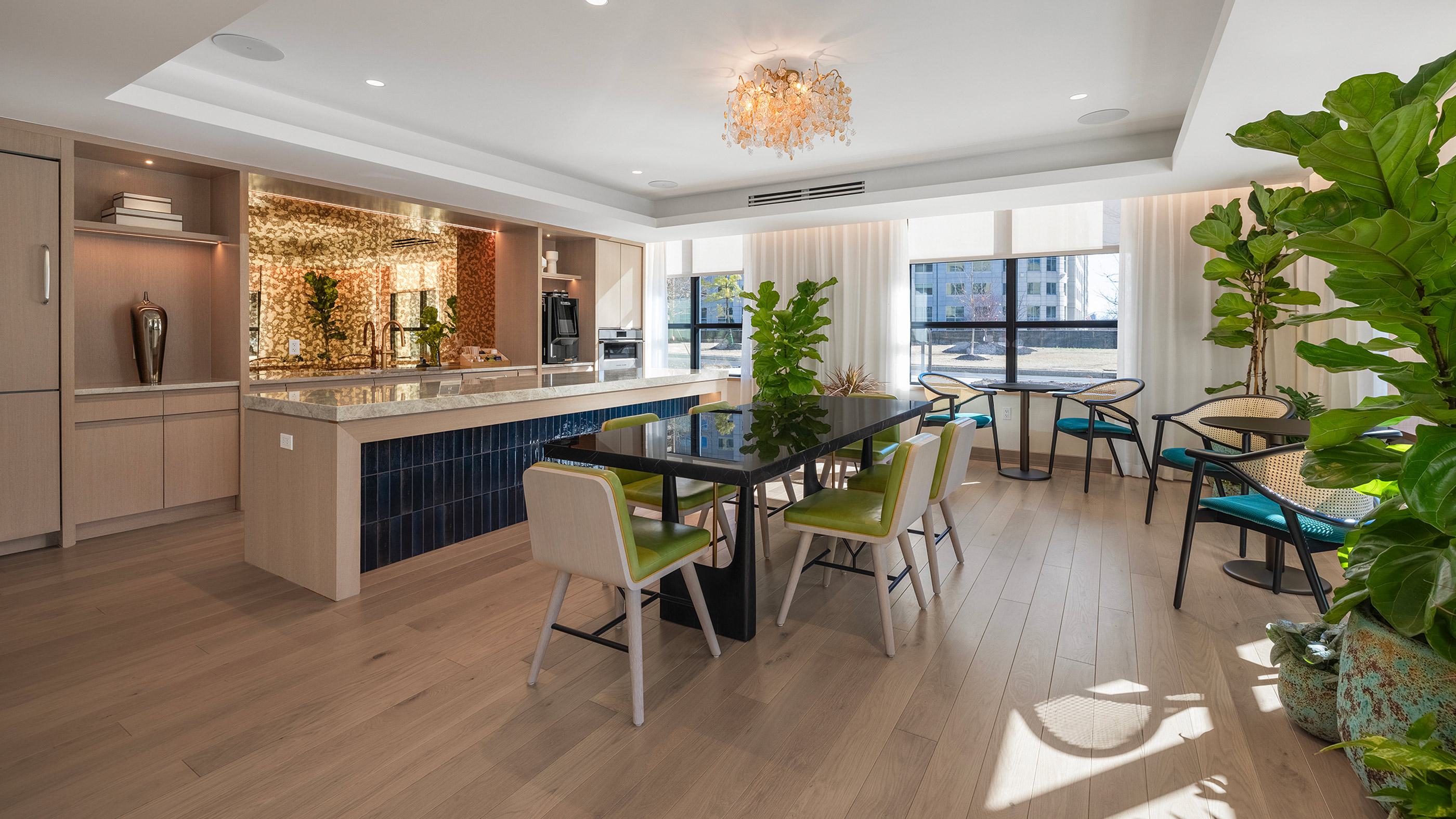 Bright orangerie-style lounge with floor-to-ceiling windows, vibrant green and teal seating, lush potted plants, a glossy black dining table, and a statement chandelier above a marble-topped serving bar with a shimmering gold backsplash.