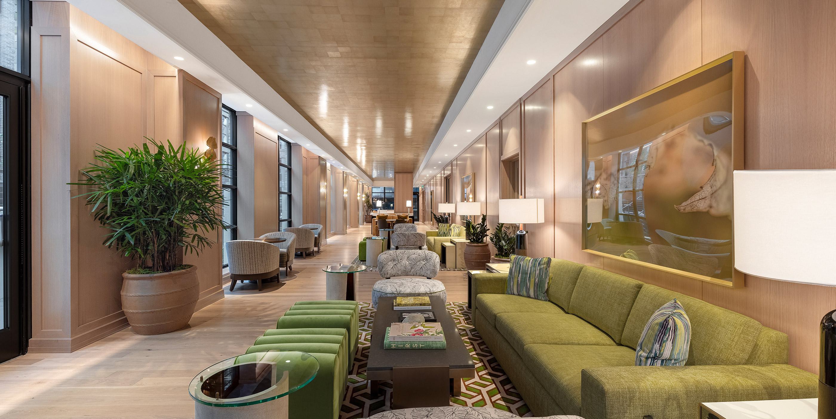 Expansive garden lounge with green and neutral-toned seating, lush potted plants, a patterned area rug, and soft wood-paneled walls, all illuminated by a golden ceiling and natural light from tall windows.