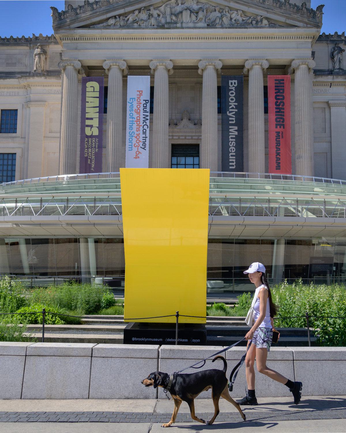 A woman walks her dog past the Brooklyn Museum, with its grand neoclassical facade, colorful exhibition banners, and a bold yellow outdoor sculpture in view.