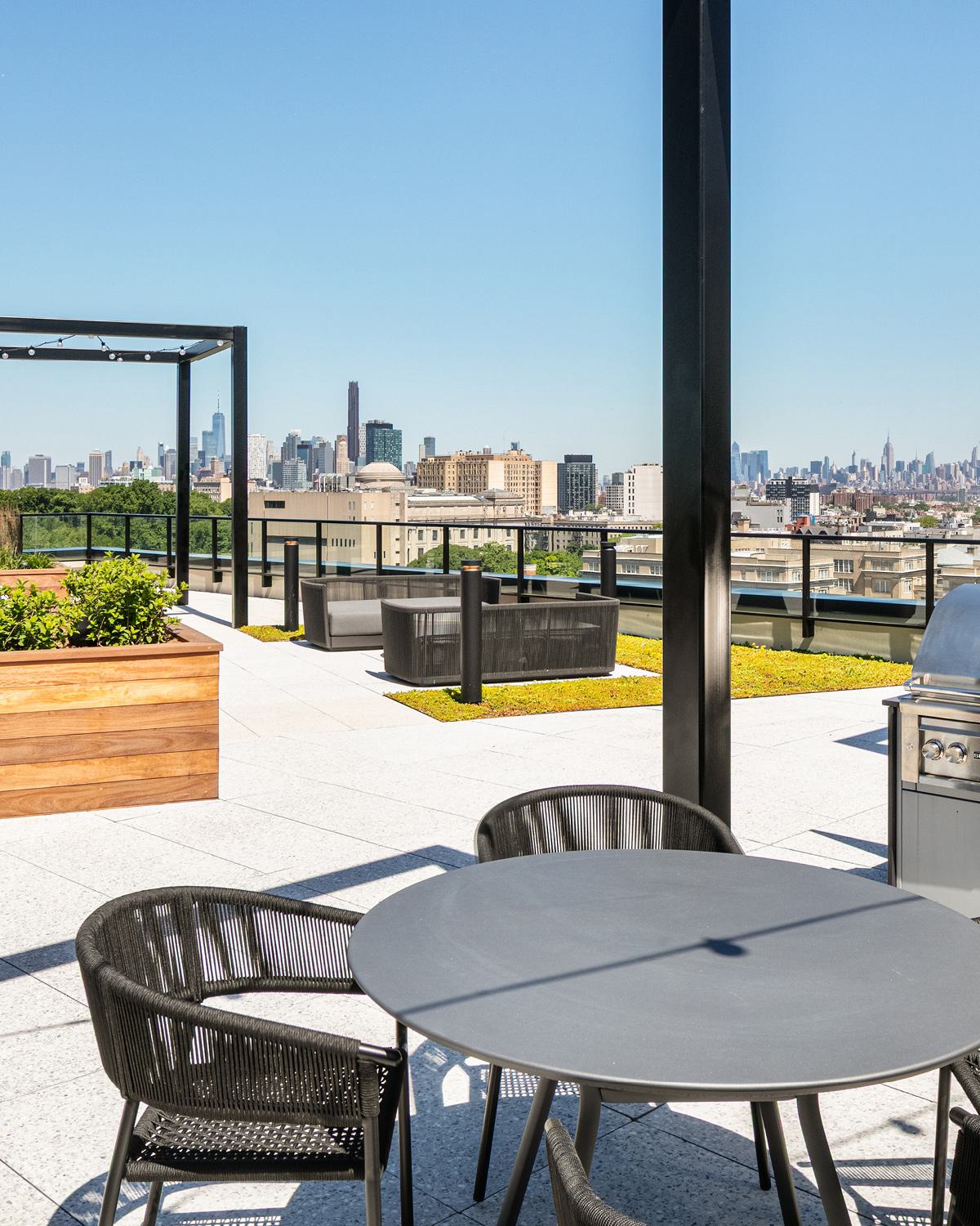 rooftop terrace with lush greenery, stylish outdoor dining, and cozy lounge seating, overlooking the Manhattan skyline at sunset with string lights above