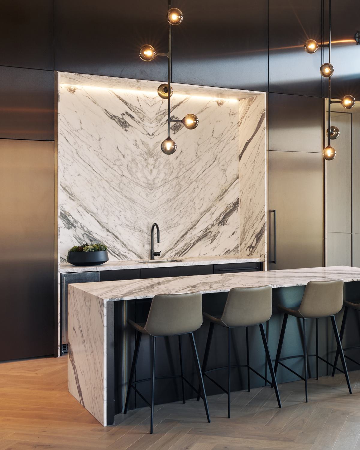 A refined lounge and dining space at Maison 78 featuring a striking waterfall marble island, brass fixtures, herringbone flooring, contemporary globe chandeliers with green glass accents, and large windows framing views of Manhattan architecture. Upholstered chairs and soft wood tones add warmth to the sophisticated interior.