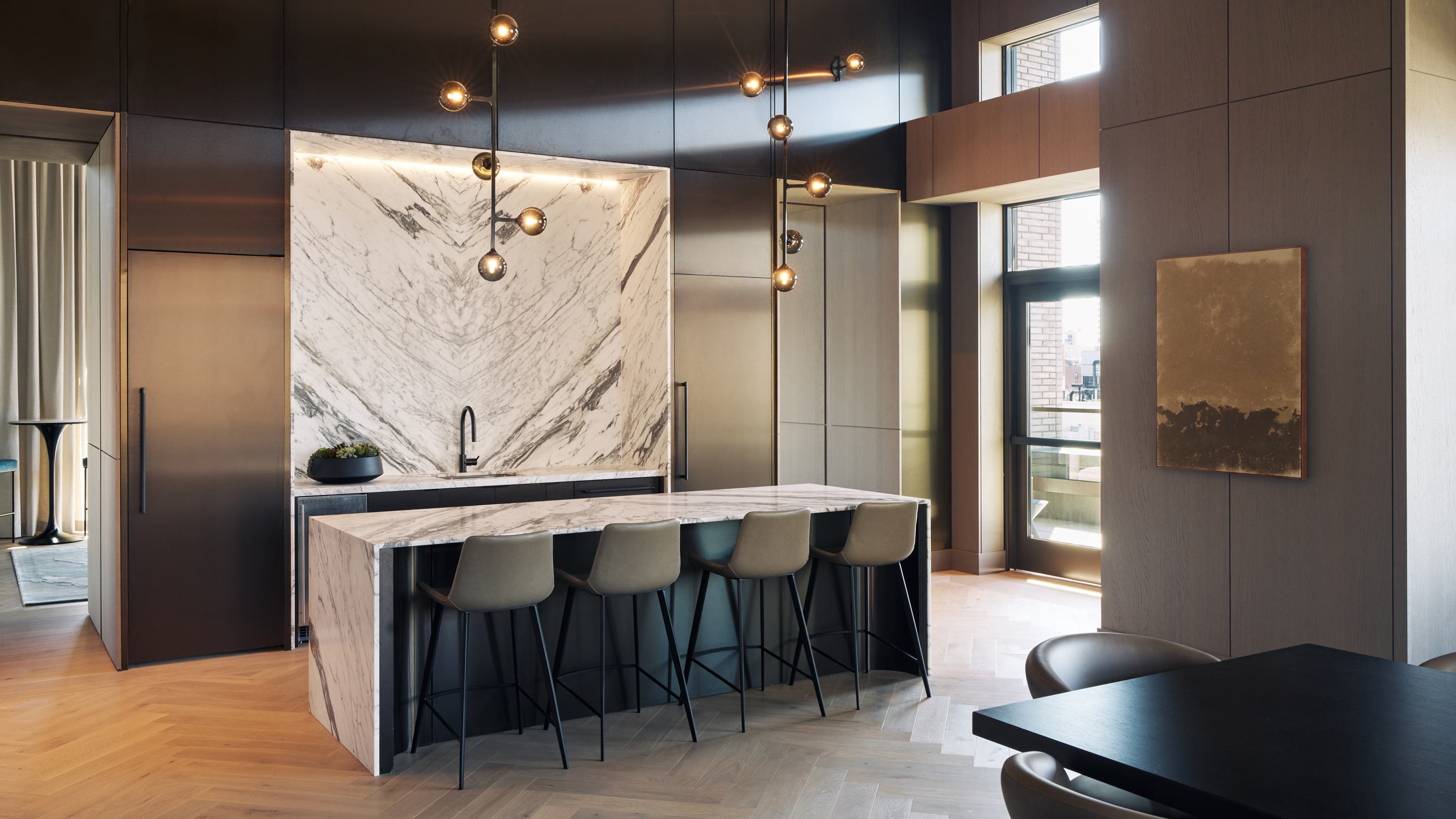 A refined lounge and dining space at Maison 78 featuring a striking waterfall marble island, brass fixtures, herringbone flooring, contemporary globe chandeliers with green glass accents, and large windows framing views of Manhattan architecture. Upholstered chairs and soft wood tones add warmth to the sophisticated interior.