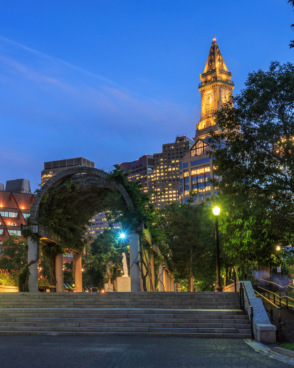 Christopher Columbus Waterfront Park in Boston, with the Boston Custom House Tower prominently visible in the background. The park, located along the waterfront in the North End, features a beautiful trellis covered in greenery, which is a well-known spot for visitors and locals alike. The Custom House Tower, an iconic Boston landmark, is illuminated in the evening,