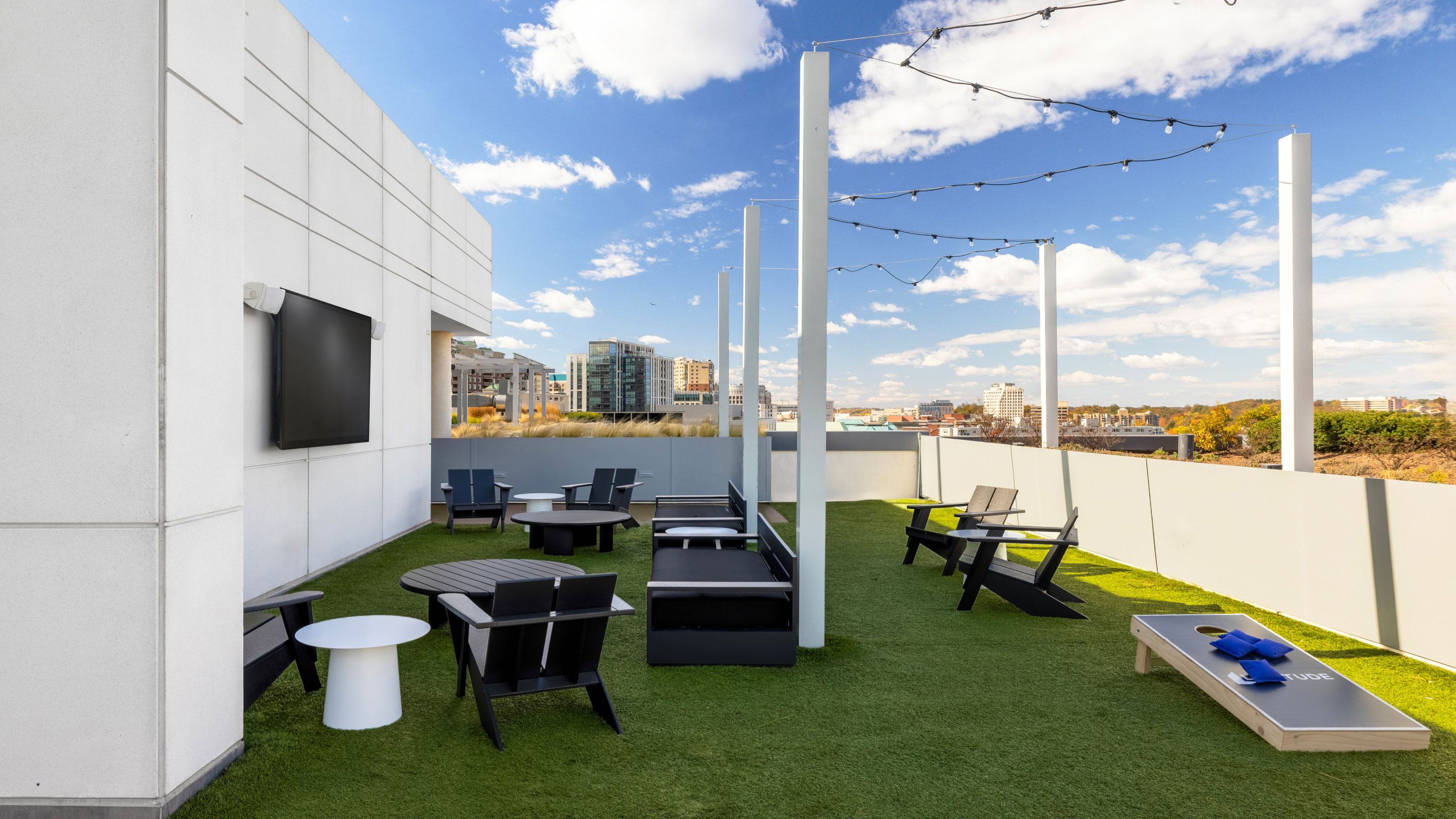 Rooftop social lawn at Latitude Apartments in Virginia Square with turf seating, outdoor lounge furniture, string lighting, yard games, and elevated views across Arlington’s tree-lined neighborhoods.