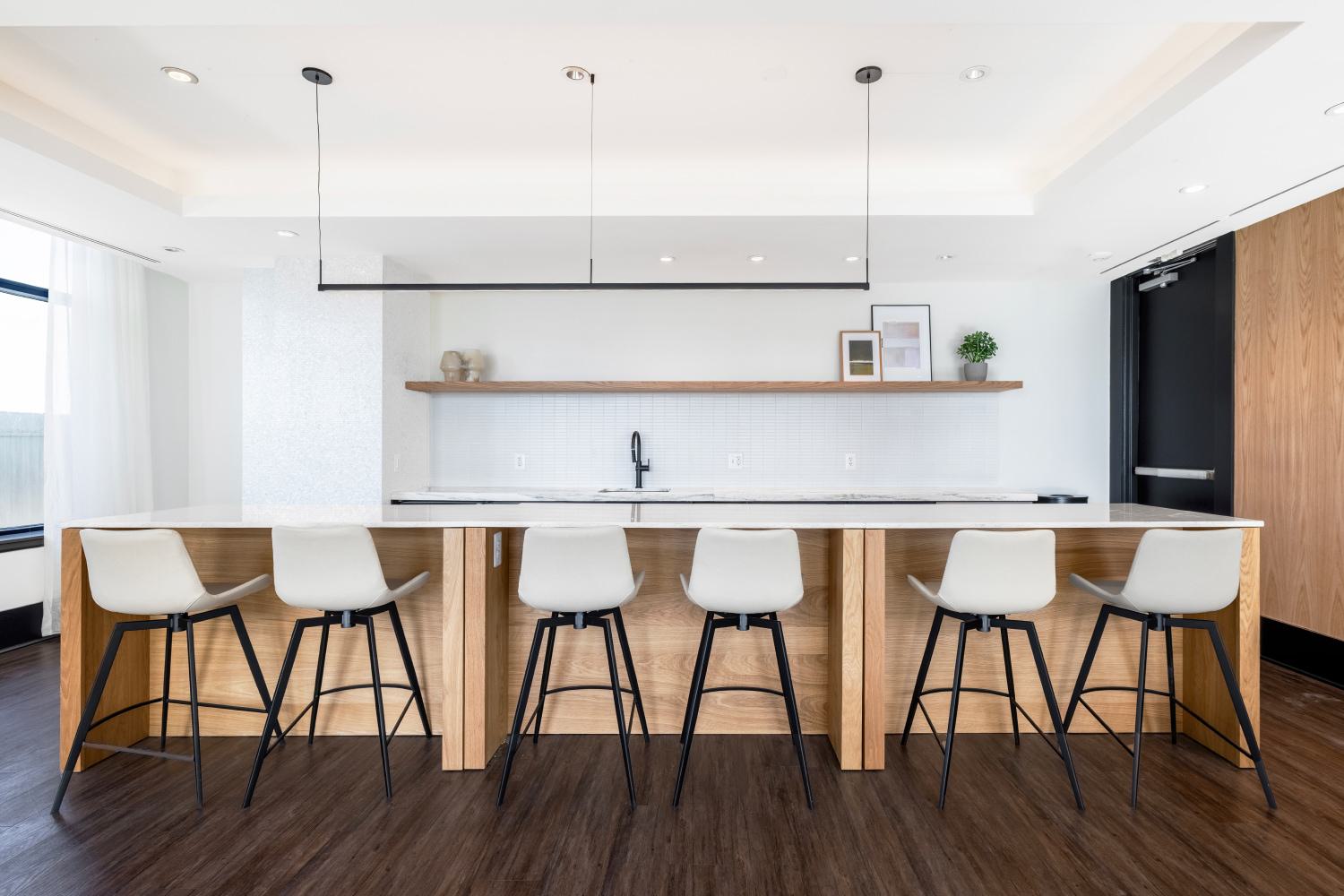 Community kitchen and bar area at Latitude Apartments in Virginia Square featuring a long wood island, modern bar seating, open shelving, and bright contemporary finishes.