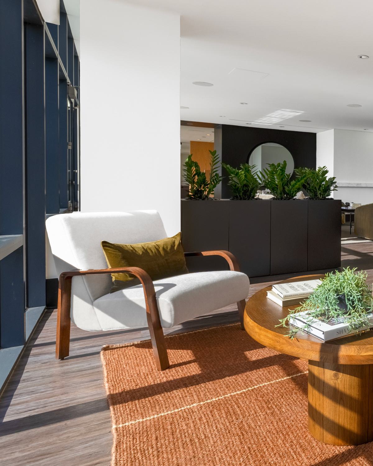 Sunlit lounge seating area at Latitude Apartments in Virginia Square with a modern white armchair, wood coffee table, indoor greenery, and warm wood flooring.