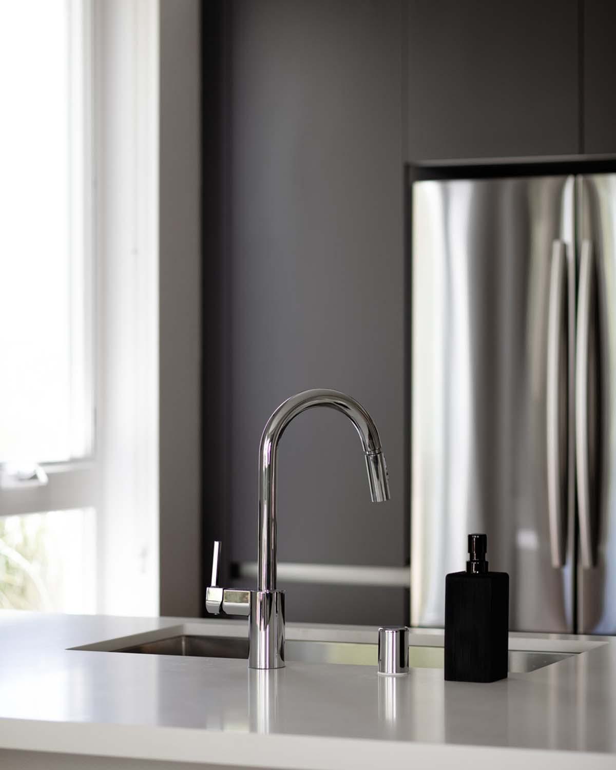 Close-up of a modern apartment kitchen island at Latitude Apartments in Arlington, VA, featuring a polished chrome gooseneck faucet set into a smooth stone countertop, with stainless steel refrigerator and minimalist cabinetry in the background.