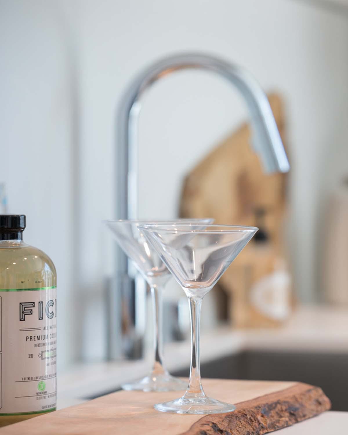 Close-up of a styled apartment kitchen sink at Latitude Apartments in Arlington, VA, featuring a chrome gooseneck faucet, integrated undermount sink, minimalist countertop accessories, and cocktail glasses arranged on a wood tray, with soft natural light in the background.
