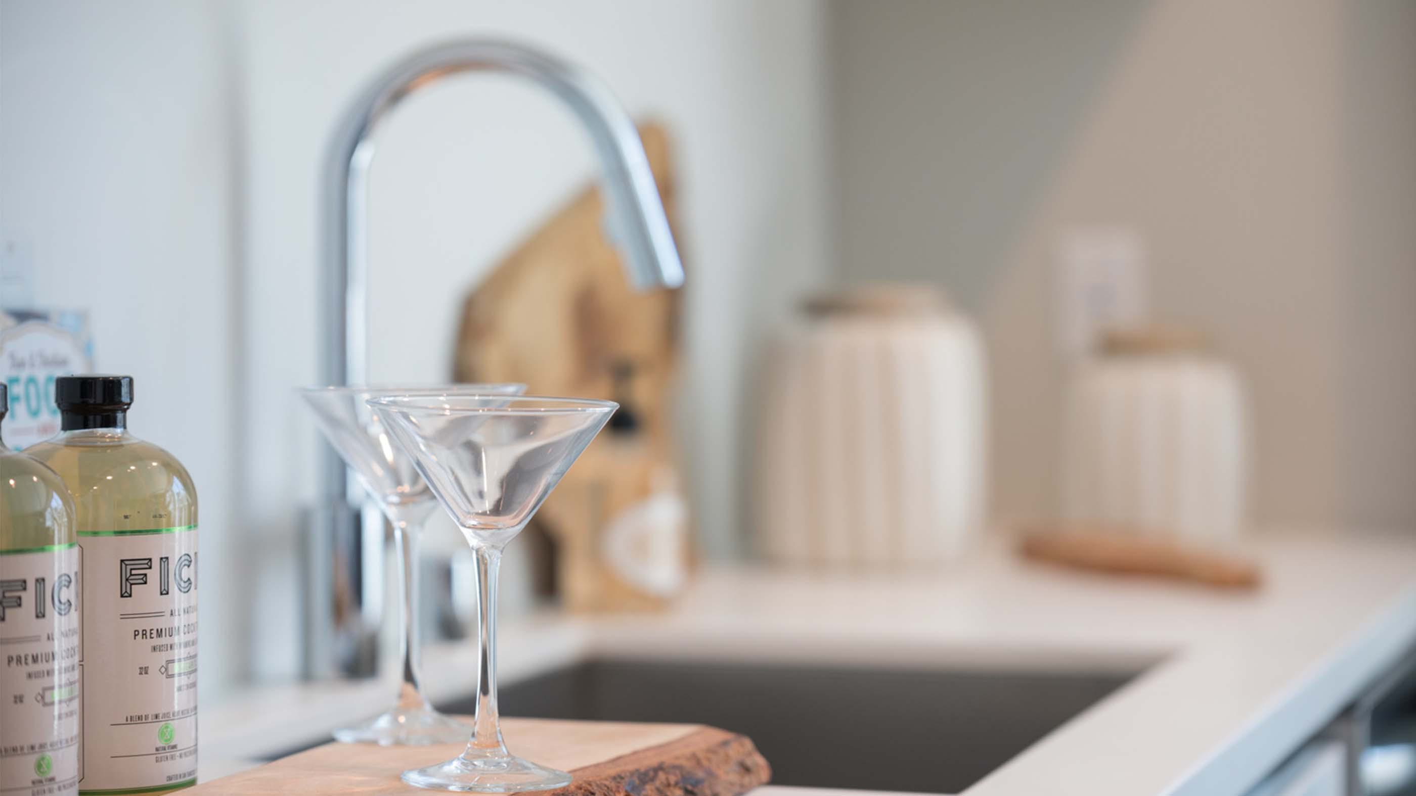 Close-up of a styled apartment kitchen sink at Latitude Apartments in Arlington, VA, featuring a chrome gooseneck faucet, integrated undermount sink, minimalist countertop accessories, and cocktail glasses arranged on a wood tray, with soft natural light in the background.