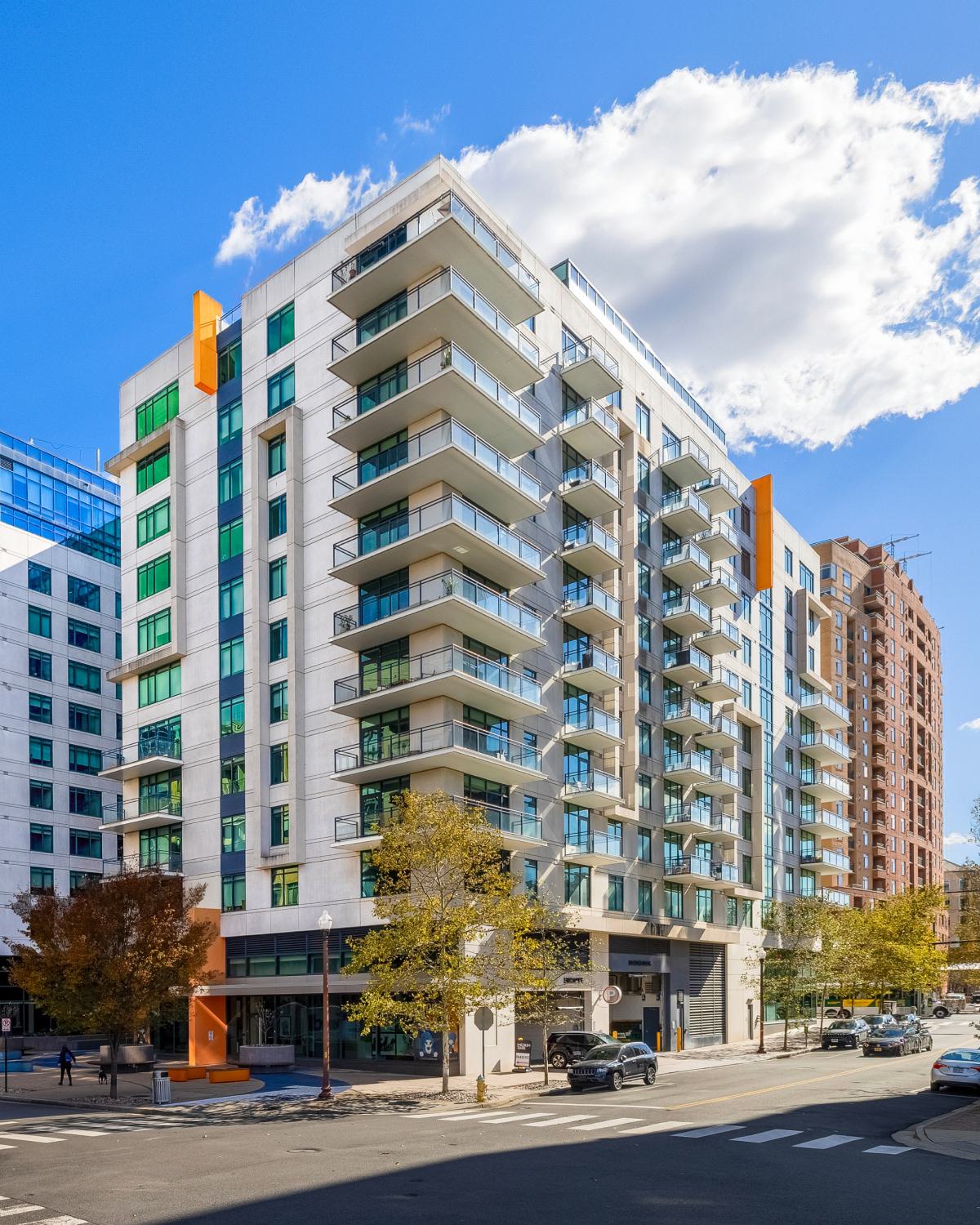 Exterior view of Latitude Apartments in Arlington, Virginia, showcasing contemporary architecture with glass balconies, street-level retail, and a prominent corner location along a tree-lined street.