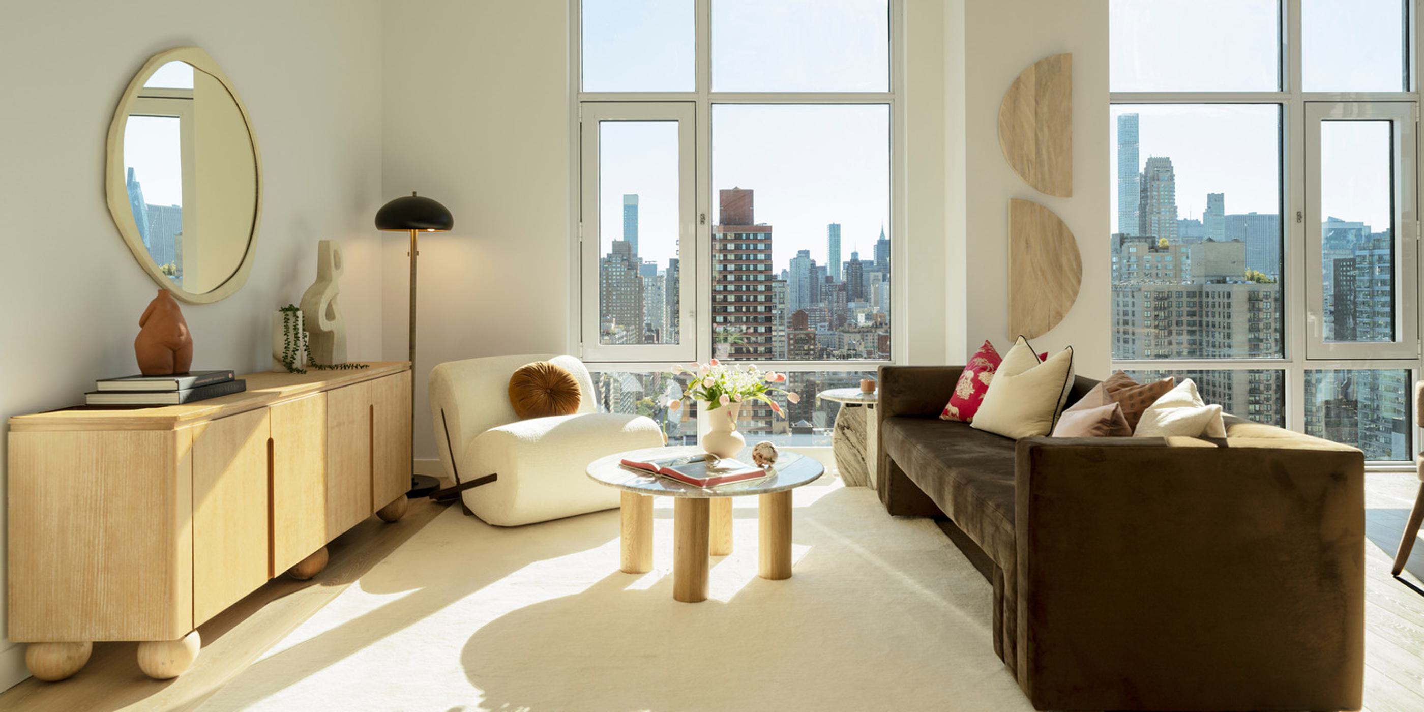 A luxurious UES penthouse living space at Maison 78 with floor-to-ceiling windows offering expansive views of Manhattan and the East River. The interior features a marble kitchen island, sculptural dining chairs, a soft curved sectional sofa, and minimalist modern décor in a neutral palette.