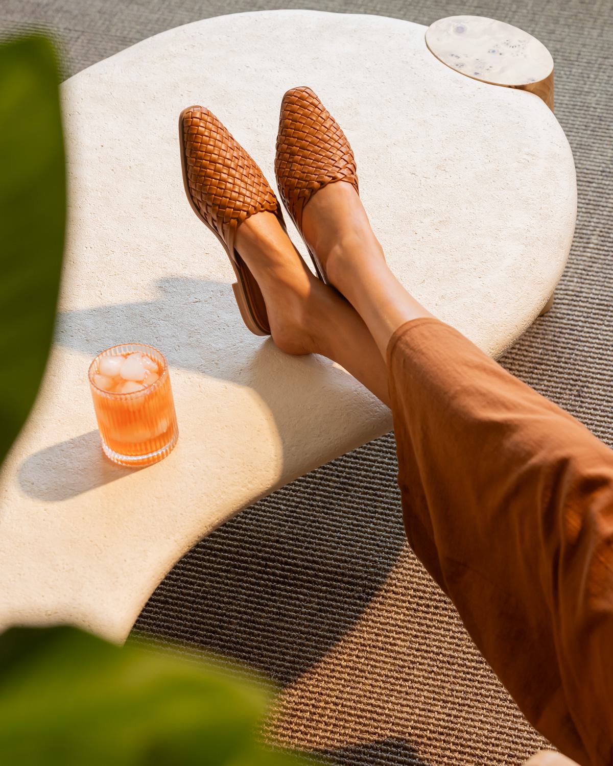 A young woman with a bright smile sits casually on a wooden bench, wearing a black top and denim jeans, surrounded by lush green monstera plants.