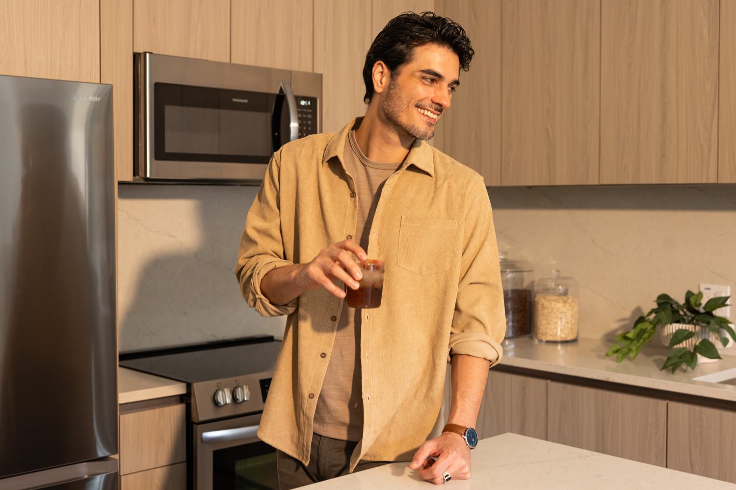 A smiling man stands in a contemporary kitchen at Rowyn apartments, holding a glass of iced coffee, surrounded by sleek cabinetry and warm natural light.