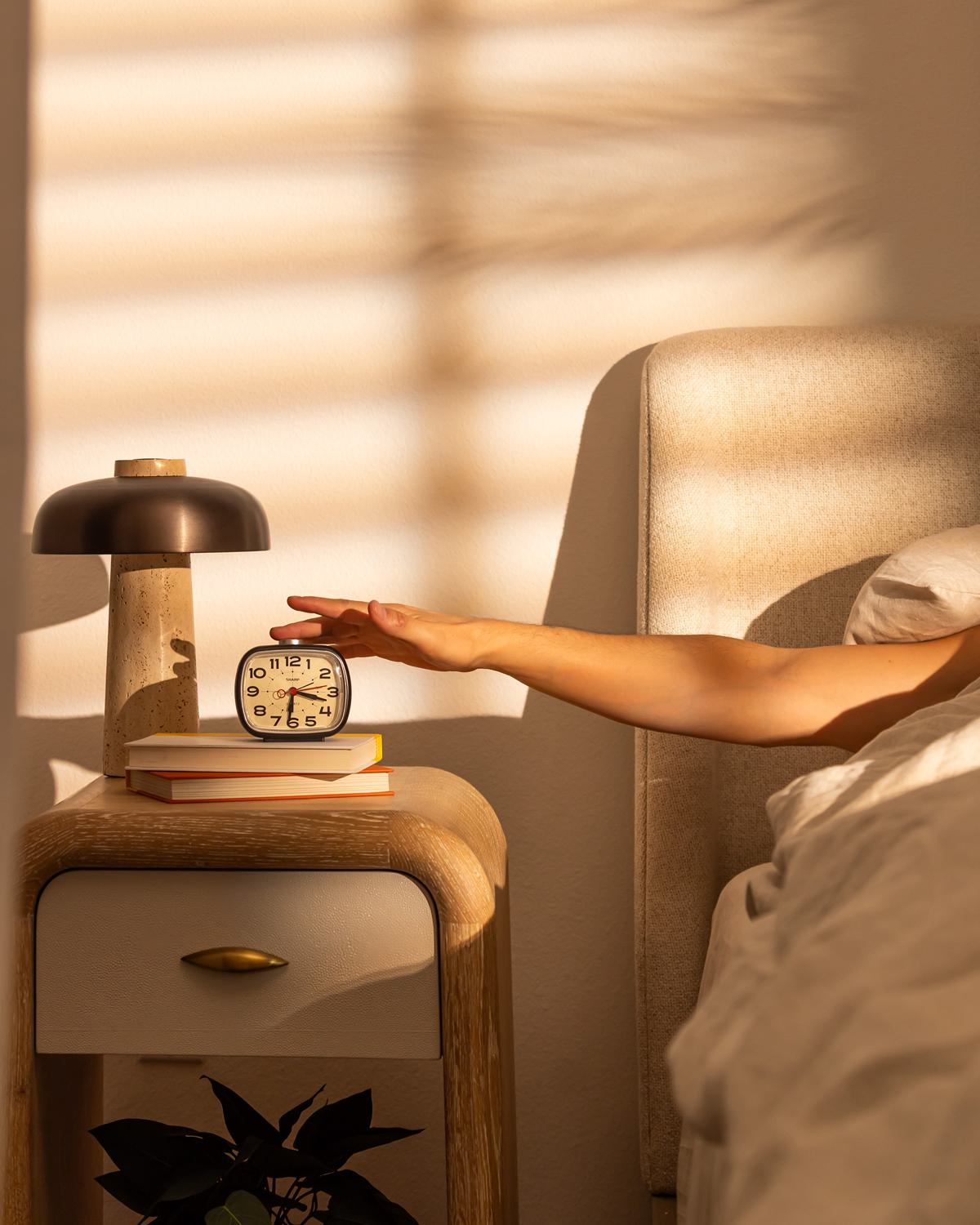 A person reaches to turn off an alarm clock on a modern nightstand, bathed in soft morning light with shadows cast through window blinds.