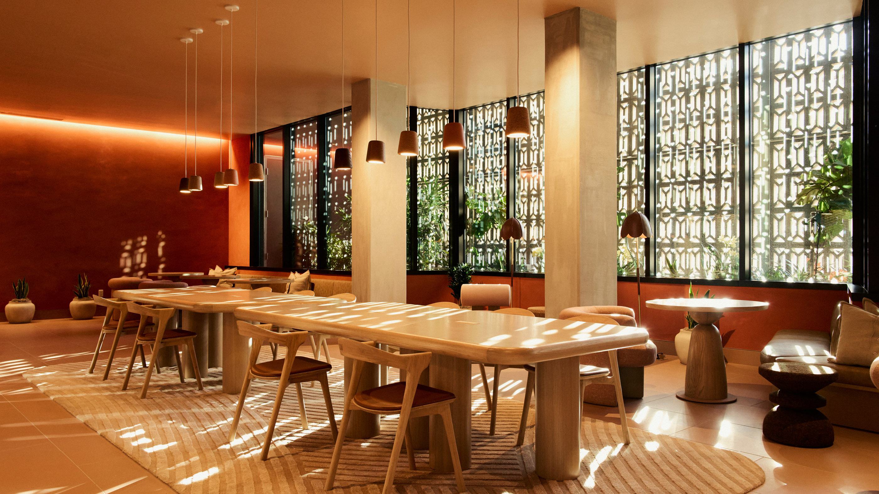 A warm, terracotta-toned lounge at Rowyn in Hillcrest, San Diego, featuring plush curved seating, soft pillows, and tiered stone tables beside a large window framing lush greenery. Built-in bookshelves line the walls, and a long wooden table with sculpted chairs and pendant lights creates a sophisticated co-working and gathering space.