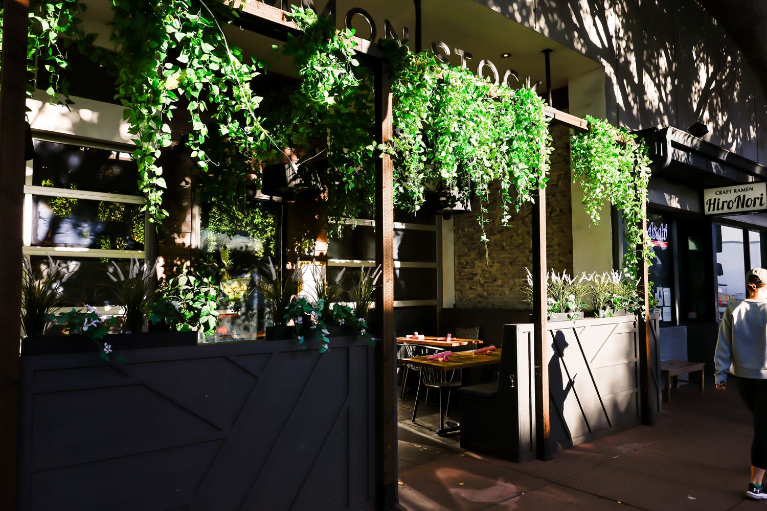 Outdoor patio of Monzu Italian Oven + Bar in San Diego’s Hillcrest neighborhood, with lush greenery, modern seating, and the neighboring HiroNori Craft Ramen visible.