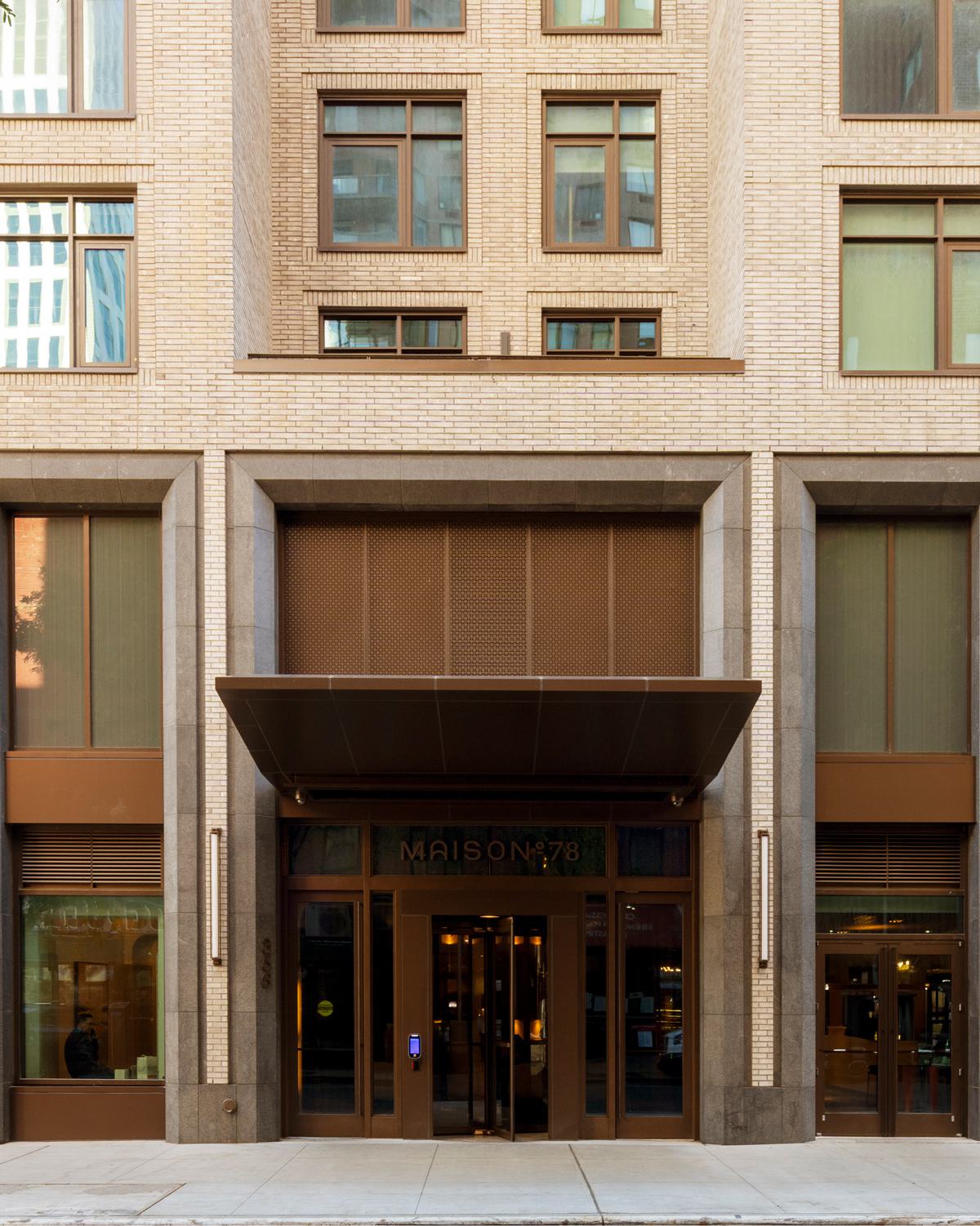 sophisticated ues building entrance with a modern facade, featuring warm lighting, elegant signage reading "MAISON°78," large glass doors, and a luxurious interior visible through the entryway, complemented by greenery and a vintage red car parked nearby.