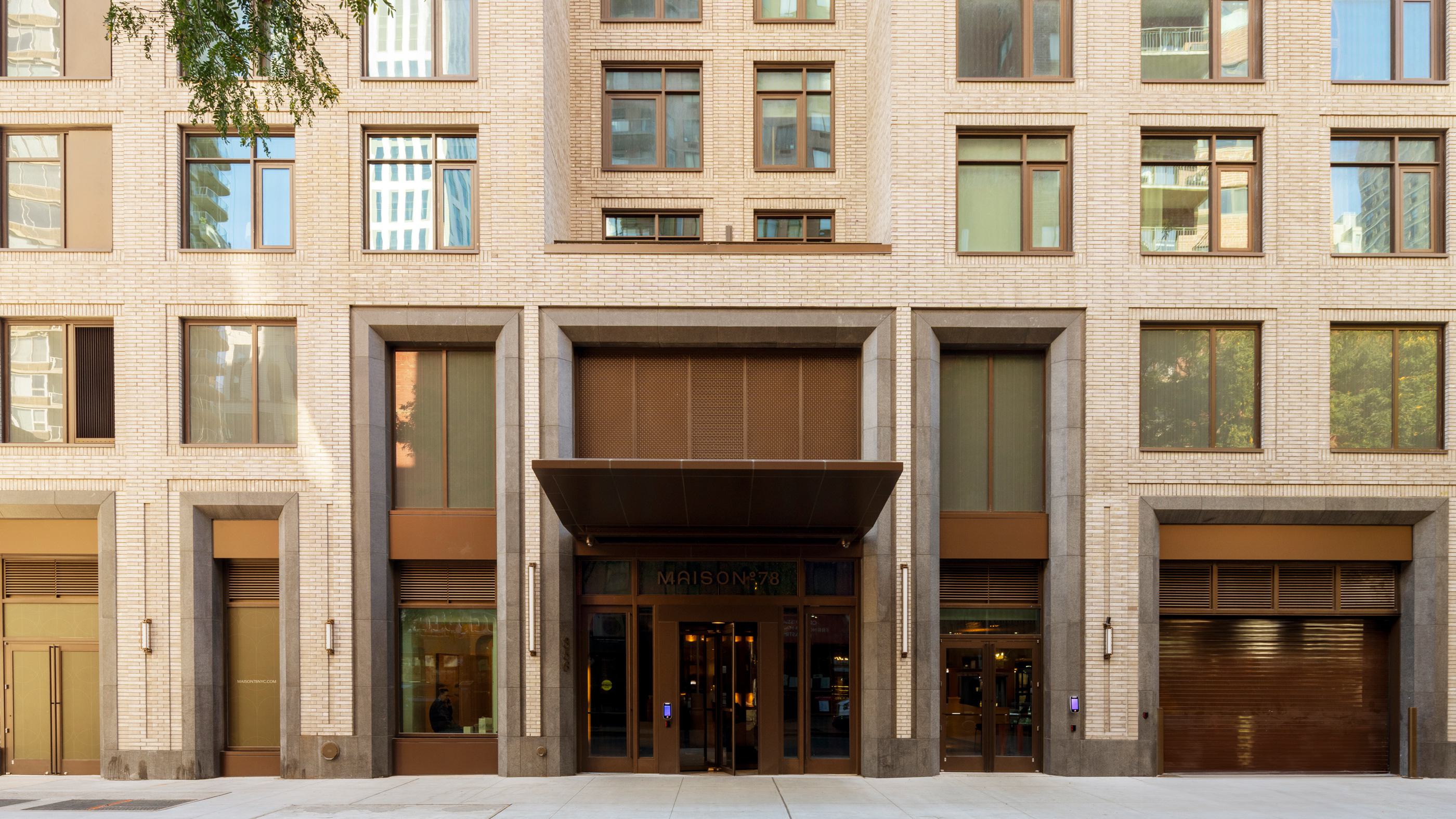sophisticated ues building entrance with a modern facade, featuring warm lighting, elegant signage reading "MAISON°78," large glass doors, and a luxurious interior visible through the entryway, complemented by greenery and a vintage red car parked nearby.