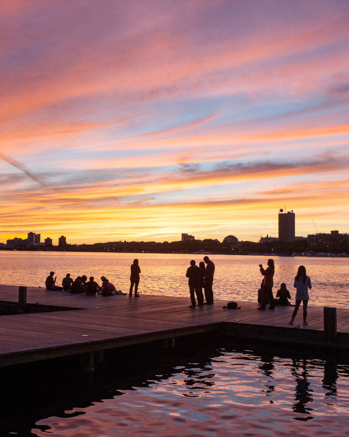 sunset over the Charles River Esplanade in Boston, with silhouetted people gathered on a wooden dock
