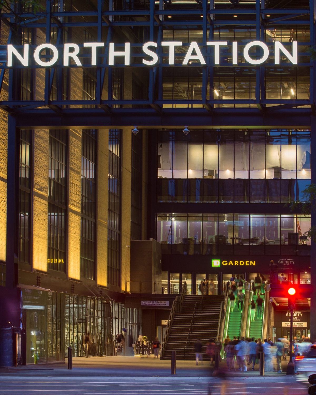 Boston’s North Station and TD Garden at night, with illuminated signage, glass architecture, and people moving up the escalators and stairs