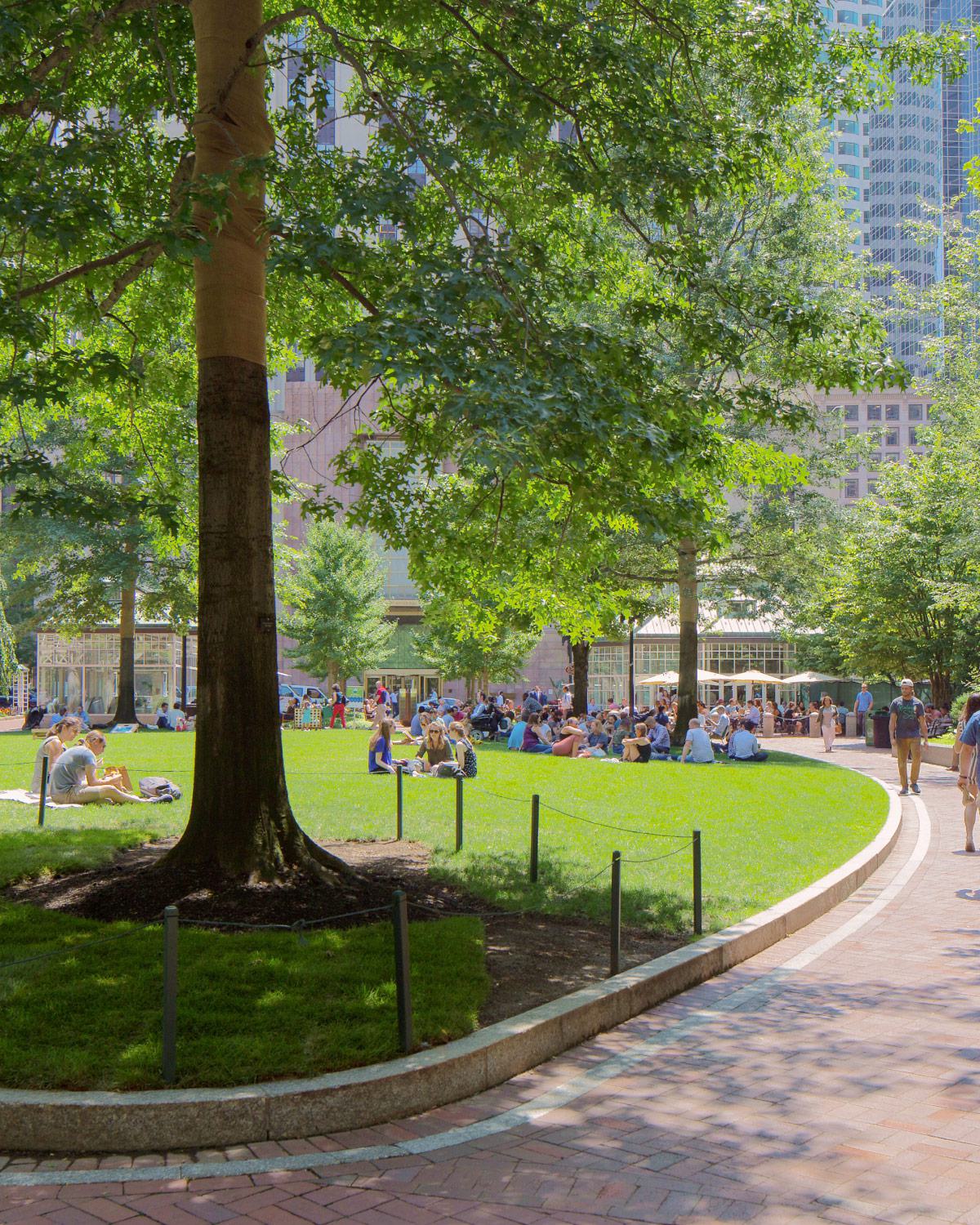 Post Office Square (Norman B. Leventhal Park) in Boston, showing a green space with people relaxing, surrounded by trees, modern office buildings, and the Glass Pavilion café in the background.