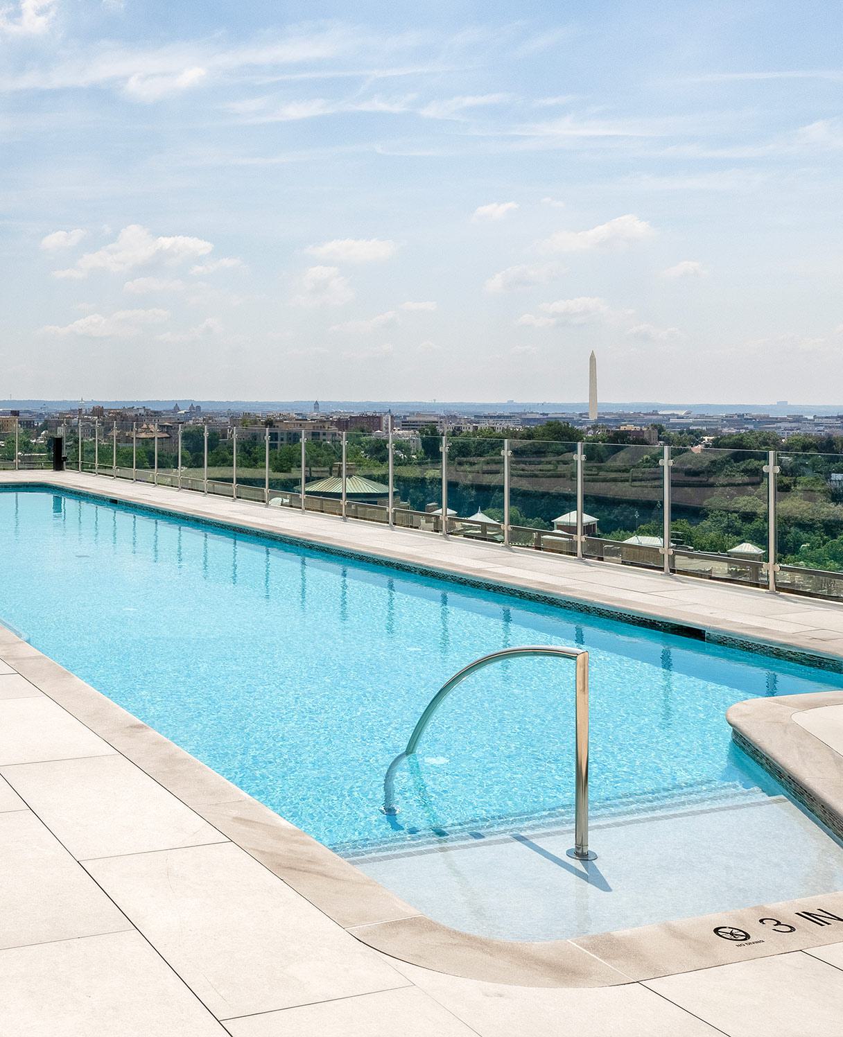 rooftop infinity pool surrounded by white lounge chairs and umbrellas, overlooking a clear blue sky.