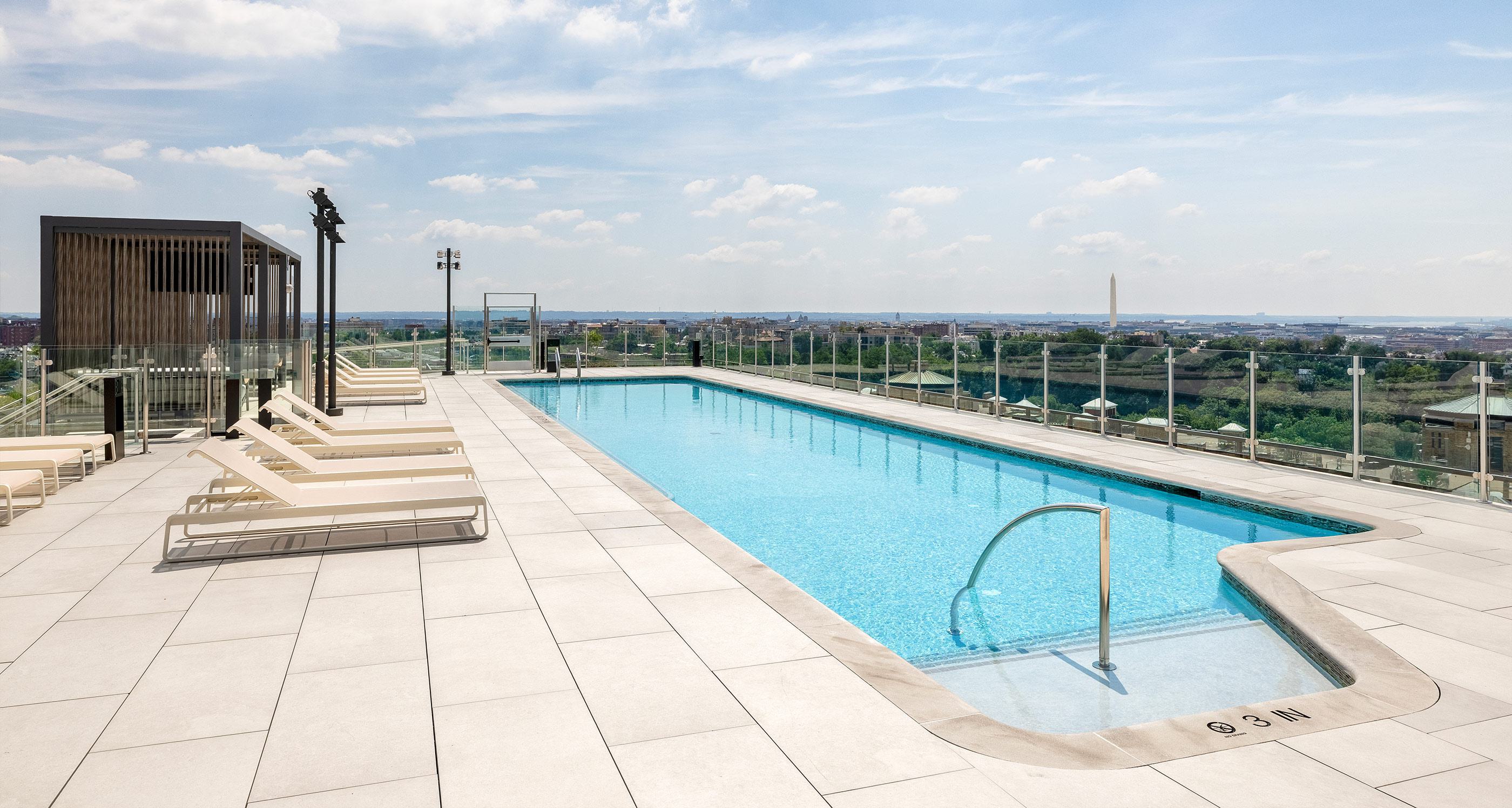 rooftop infinity pool surrounded by white lounge chairs and umbrellas, overlooking a clear blue sky.
