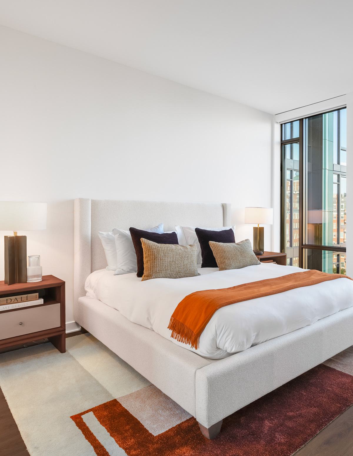 modern bedroom with floor-to-ceiling windows, and warm wood furnishings, offering a cityscape view beyond the glass.

