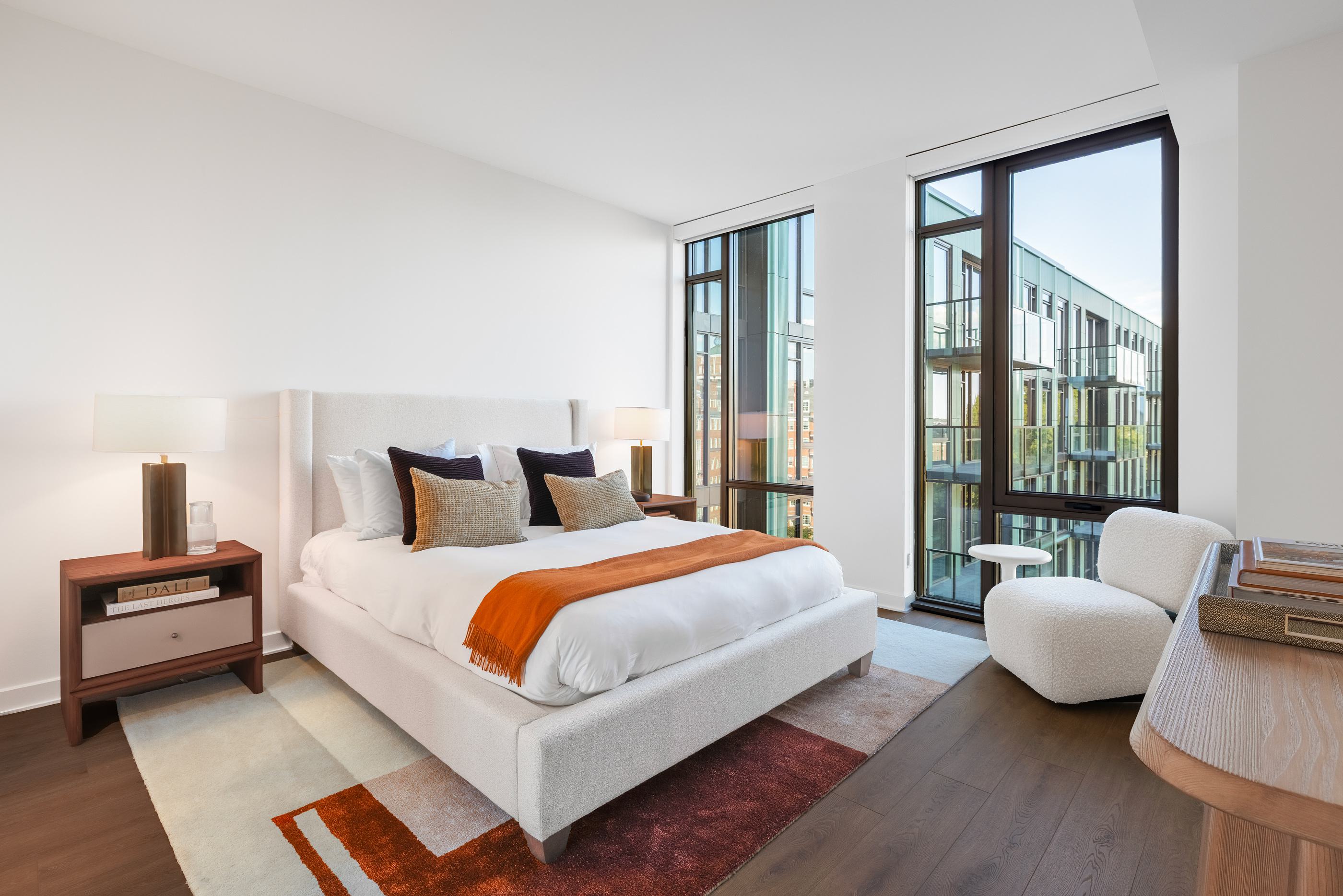modern bedroom with floor-to-ceiling windows and warm wood furnishings, offering a cityscape view beyond the glass.

