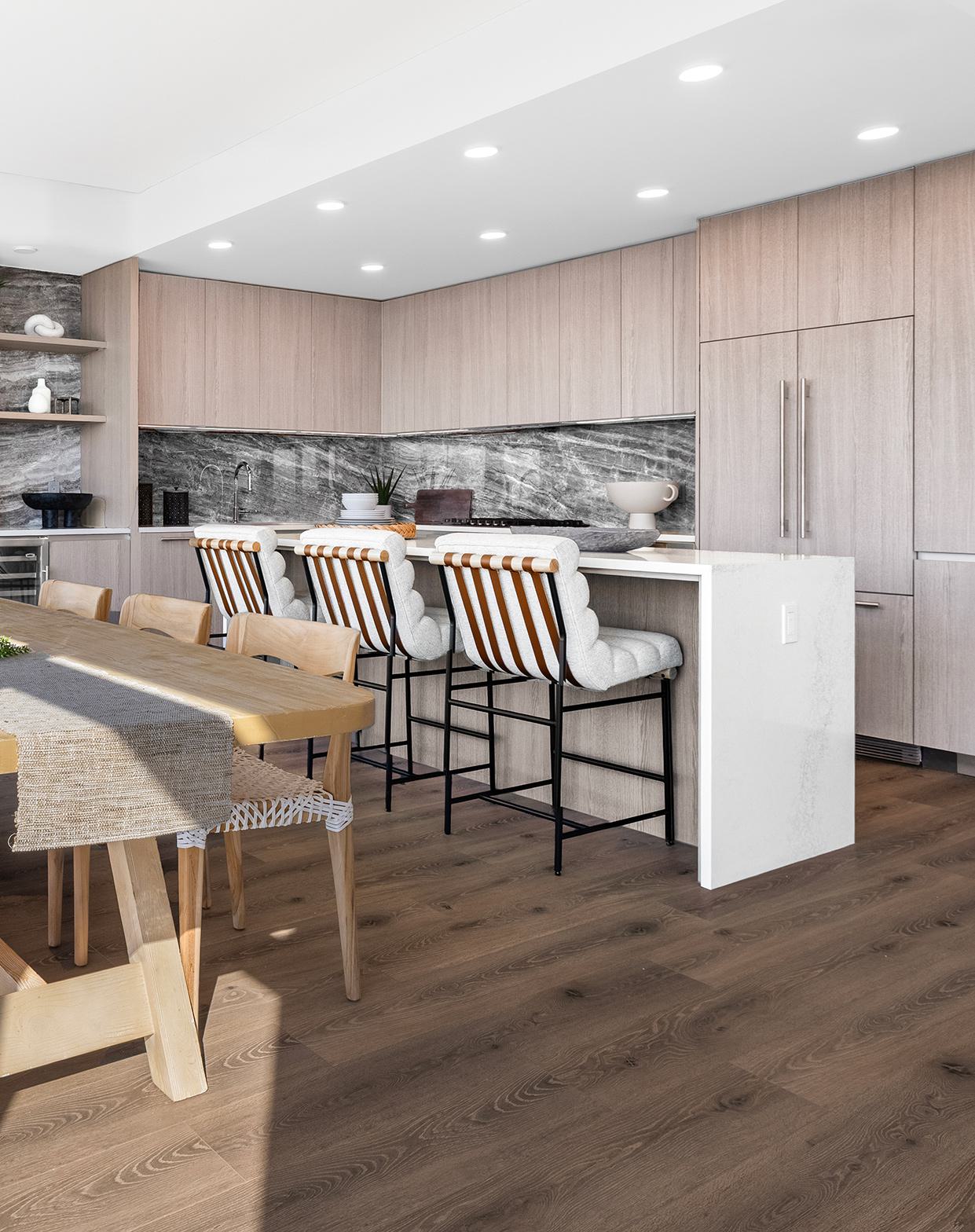 Large modern kitchen with sleek wood cabinetry, a marble waterfall island, leather barstools, and a cozy dining nook with stylish armchairs.