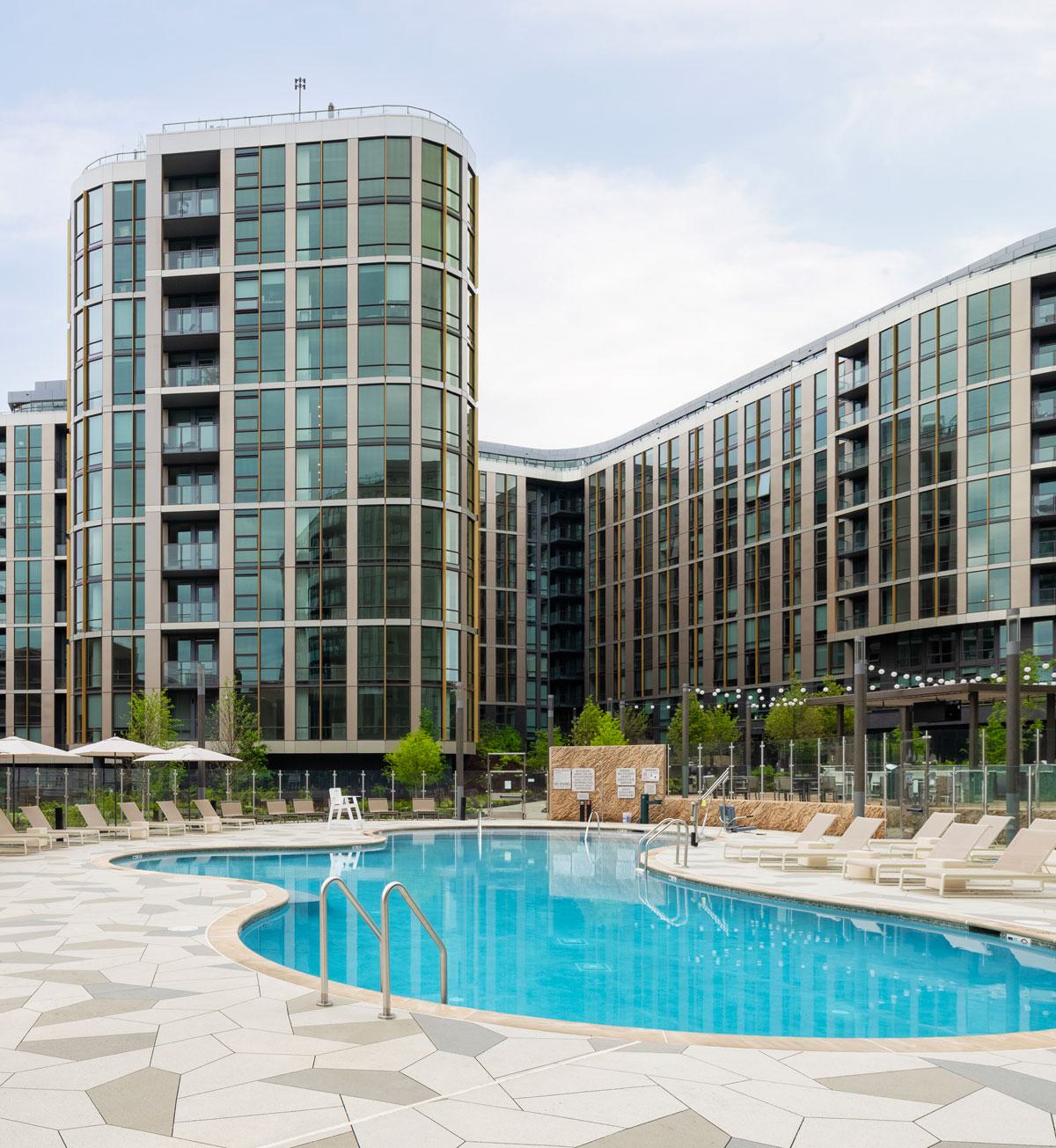 Resort-style pool at Zephyr in Woodley Park, DC, surrounded by sleek lounge chairs, umbrellas, and landscaped greenery, with the modern glass-and-metal façade of the high-rise building in the background.