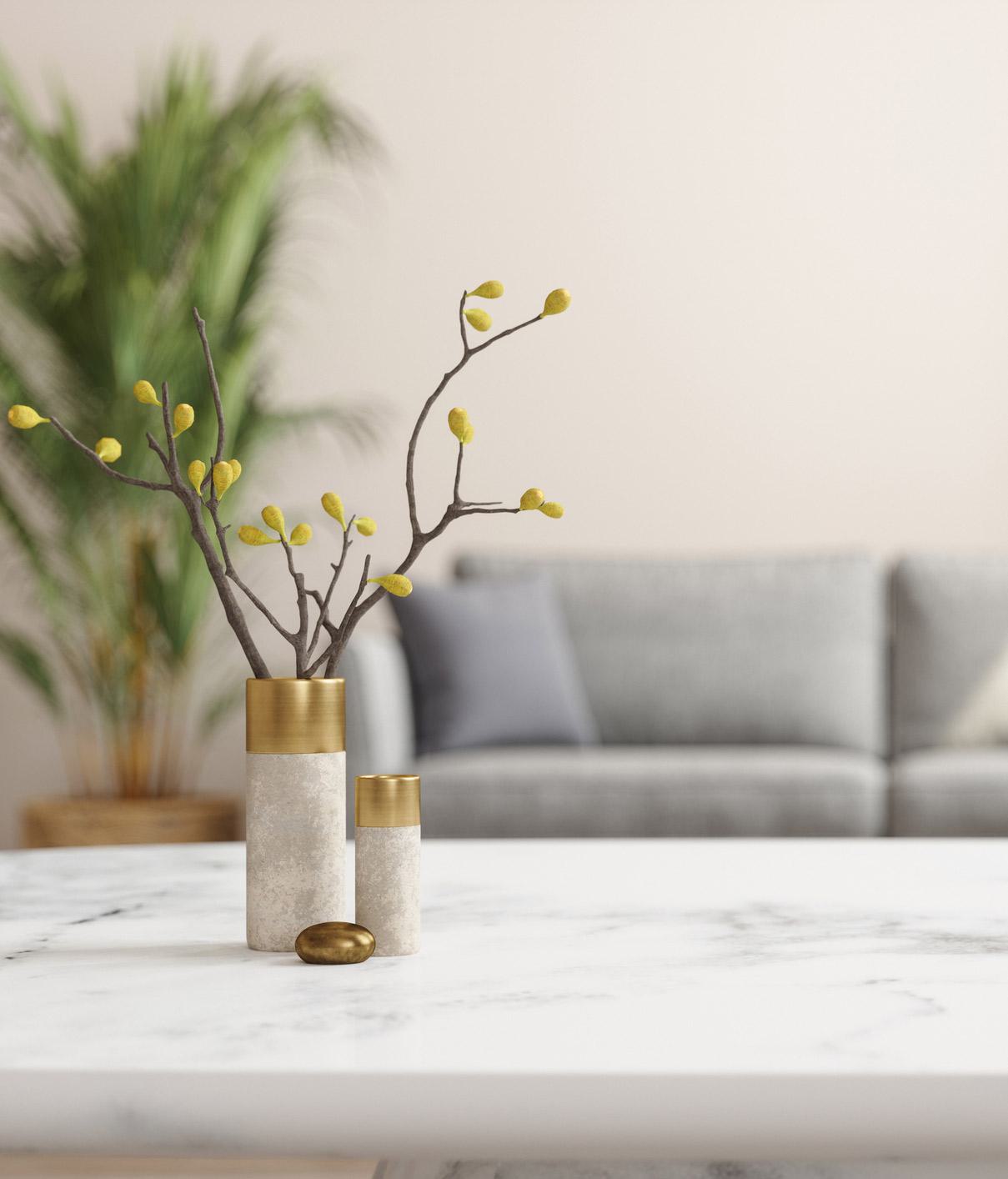 Close-up of a modern marble coffee table with two minimalist stone vases featuring brass accents and decorative branches with yellow buds, set against a blurred backdrop of a neutral-toned living room with a gray sofa and green potted plant.