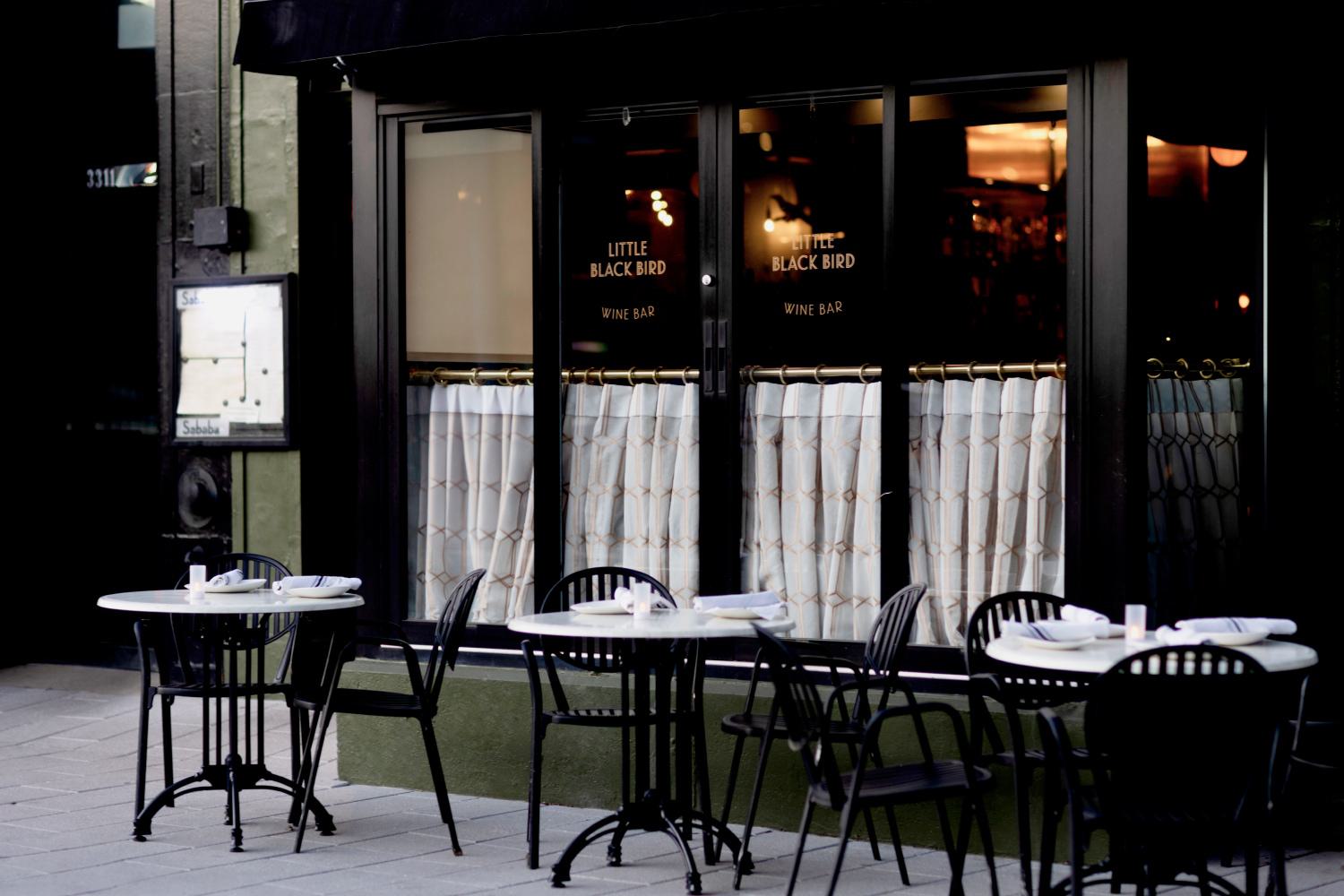 Outdoor tables set at Little Black Bird Wine Bar in Woodley Park, DC, with black chairs, white marble tops, and a cozy, intimate ambiance framed by dark-paneled windows with half curtains.