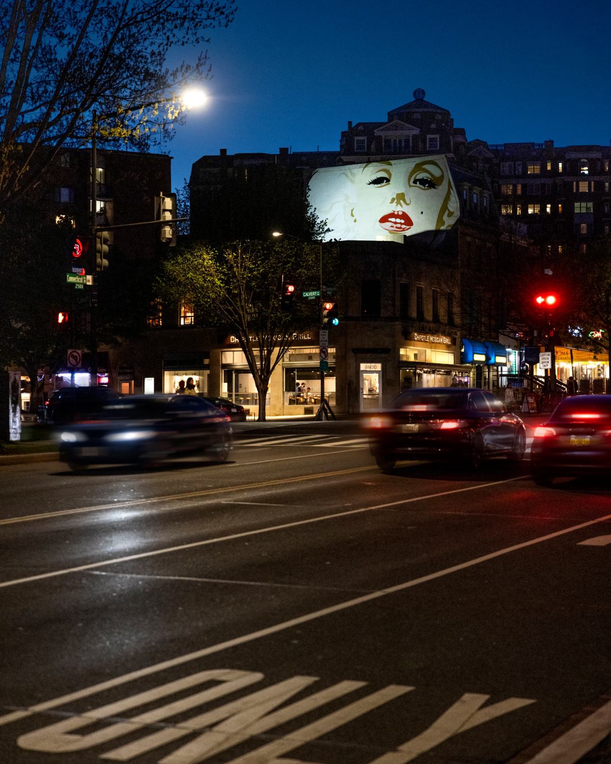 Nighttime street view in Woodley Park, DC, featuring a large illuminated mural of Marilyn Monroe above neighborhood shops and restaurants, with cars and traffic lights adding city energy to the scene.