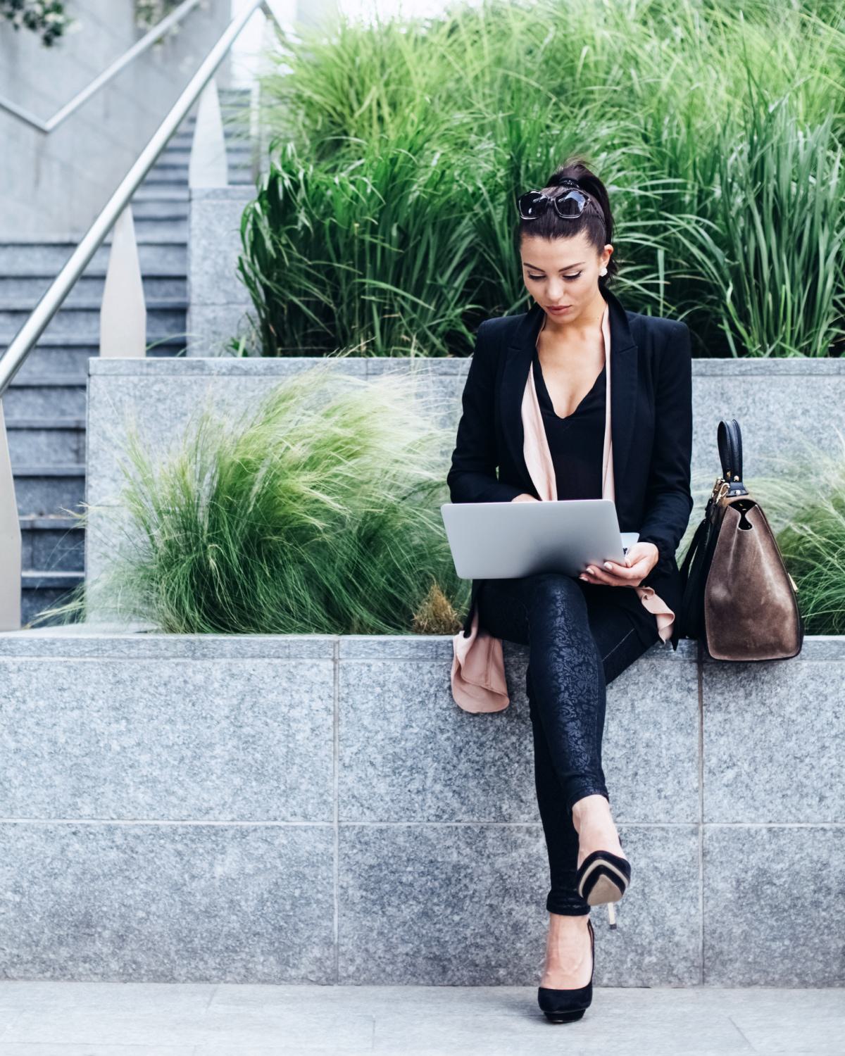 Professional woman working on a laptop outdoor,  seated on a sleek stone ledge with lush greenery in the background, representing the vibrant professional community and employers in Northwest DC.