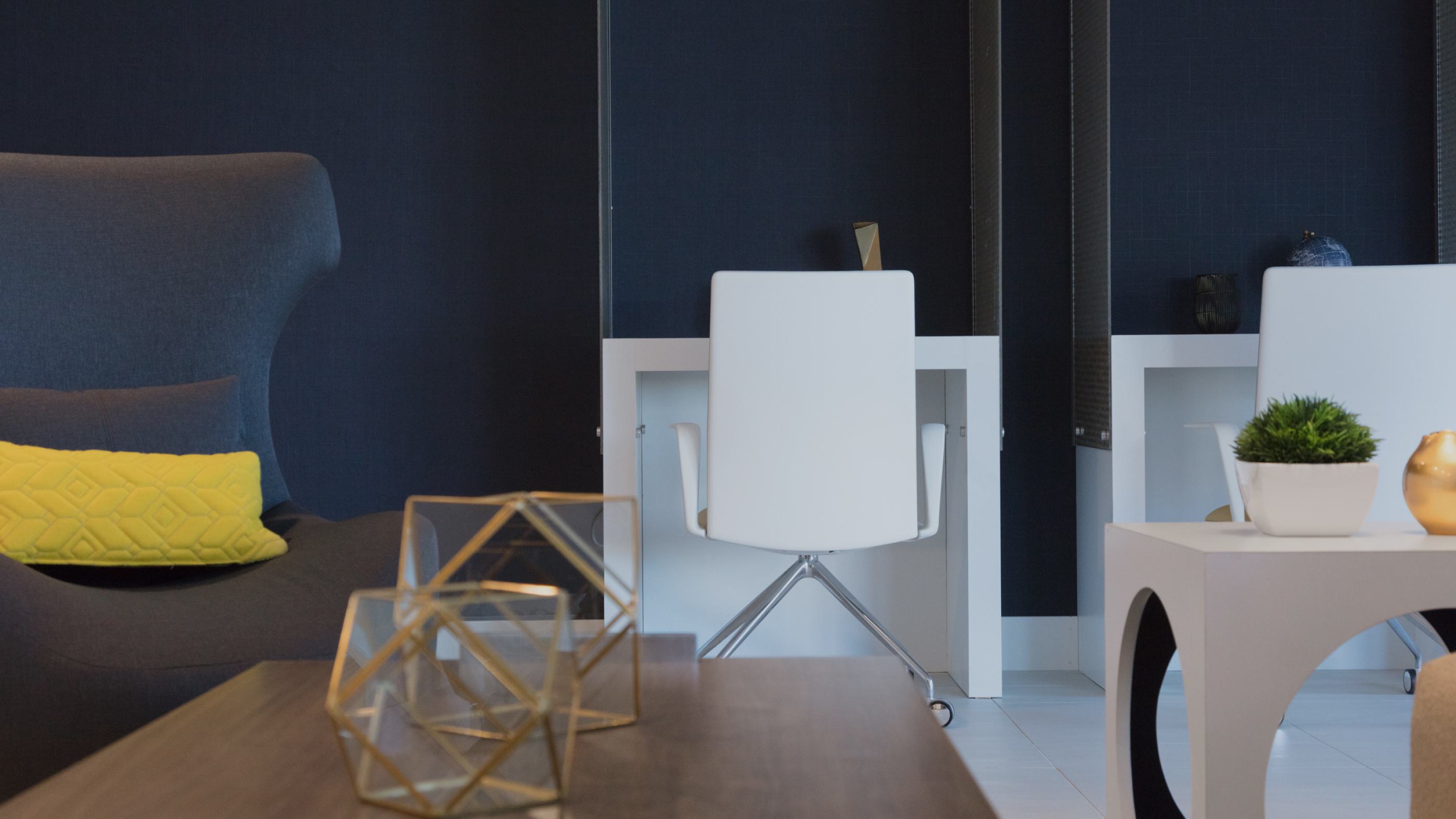 Cozy lounge and work area at The Triton Apartments featuring a modern white desk with chair, navy accent wall, geometric decor, and comfortable seating with a yellow pillow.