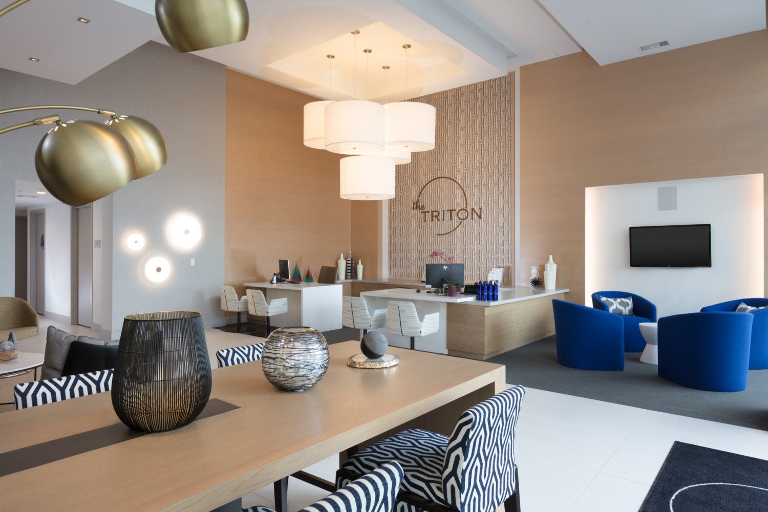 Elegant lounge at The Triton Apartments featuring modern decor, a wood accent wall with The Triton logo, statement lighting, stylish seating in blue and patterned chairs, and a welcoming reception desk.