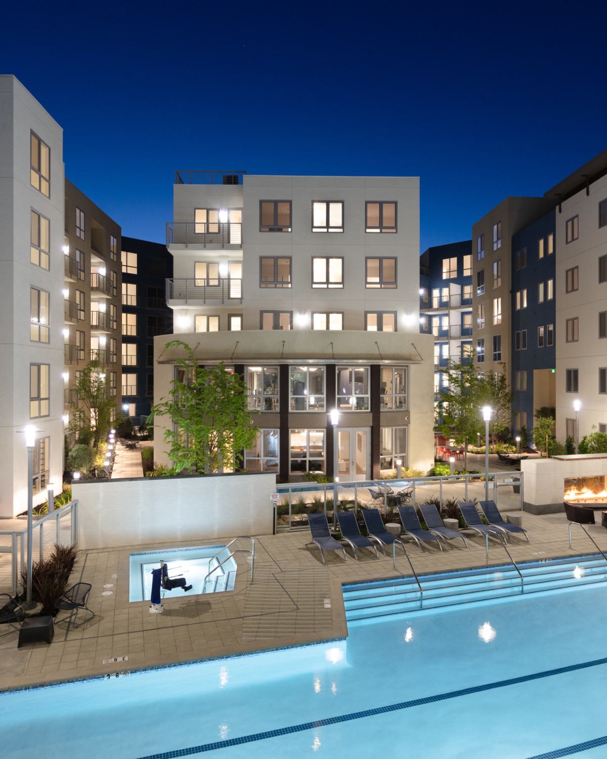 Evening view of The Triton Apartments courtyard showcasing a large illuminated swimming pool, hot tub, lounge chairs, firepit seating area, and modern apartment buildings with balconies and glowing windows.
