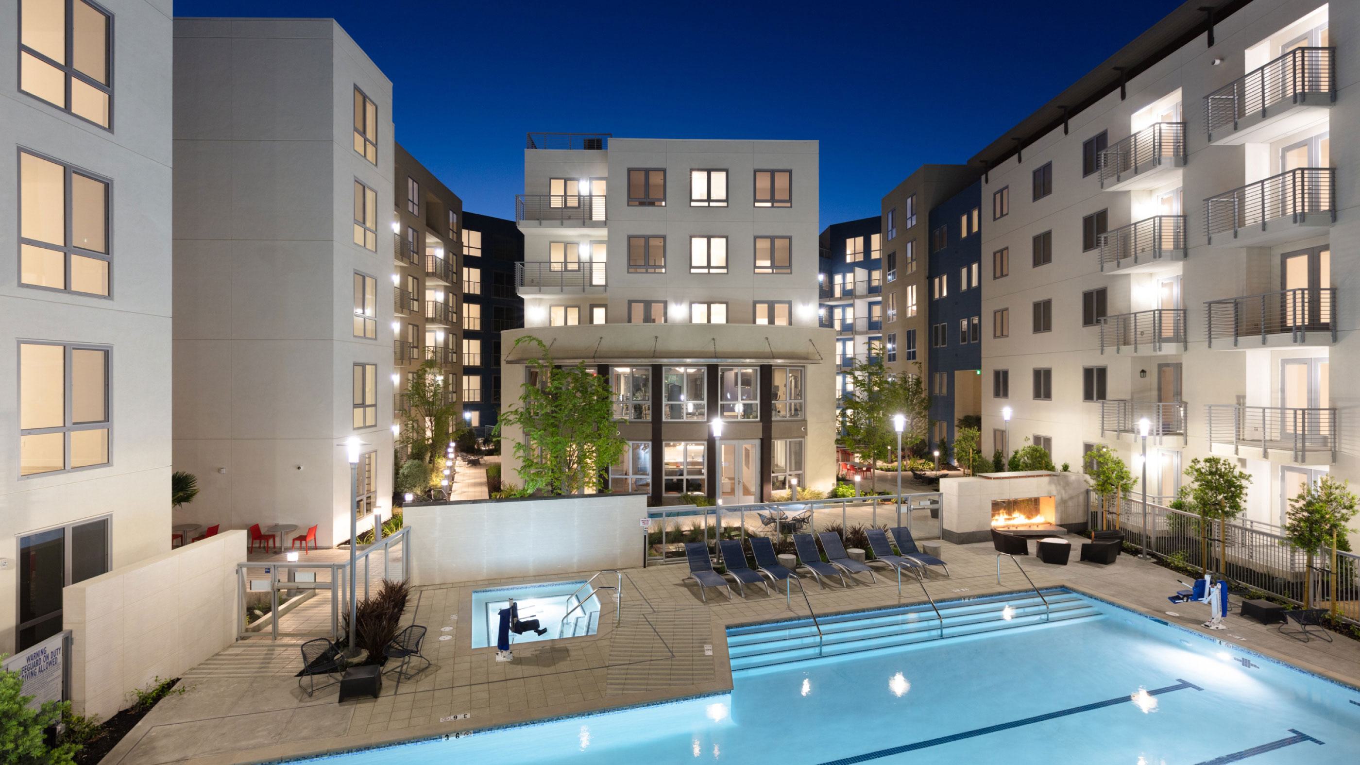 Evening view of The Triton Apartments courtyard showcasing a large illuminated swimming pool, hot tub, lounge chairs, firepit seating area, and modern apartment buildings with balconies and glowing windows.