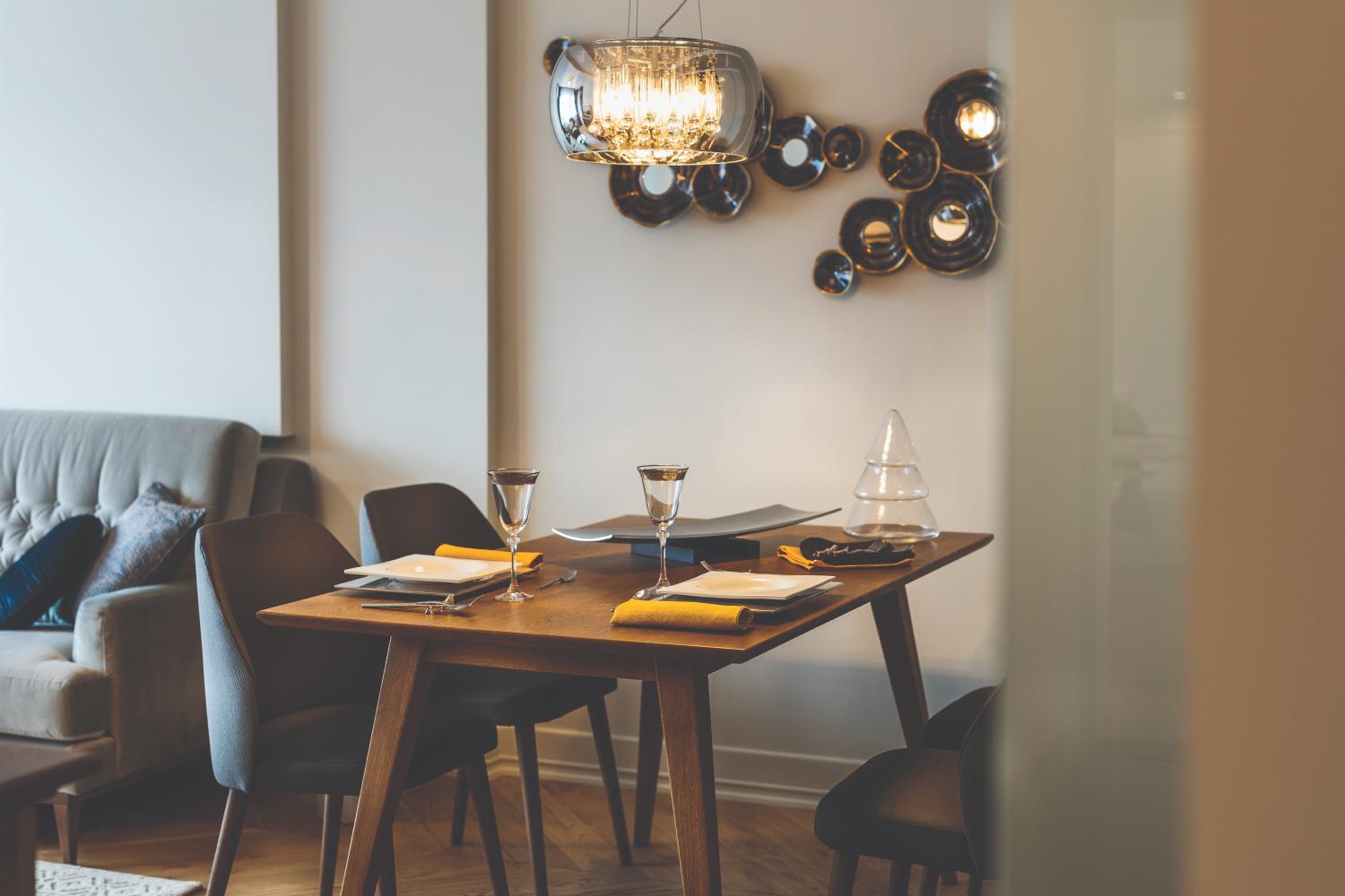 Stylish dining area with a wooden table set for two, modern gray chairs, a tufted sofa in the background, and a sculptural light fixture with decorative wall accents.