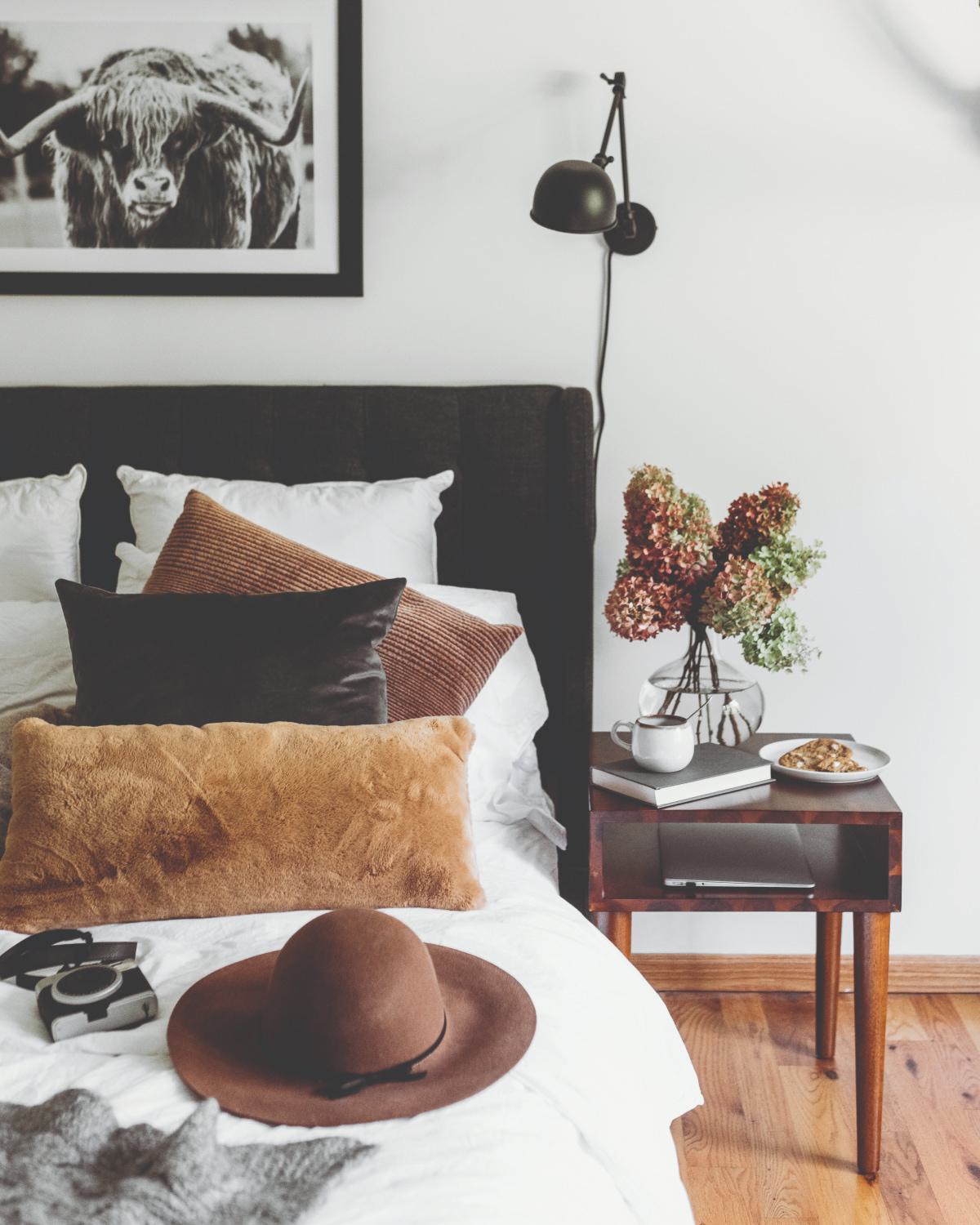 Cozy bedroom with a dark upholstered headboard, layered neutral pillows, and a bedside table styled with flowers, a book, coffee, and a pastry, creating a warm and inviting atmosphere.