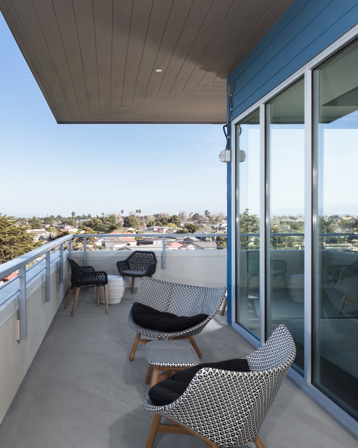 Spacious private balcony at The Triton Apartments with modern woven lounge chairs and a view overlooking treetops and neighborhood rooftops under a clear sky.
