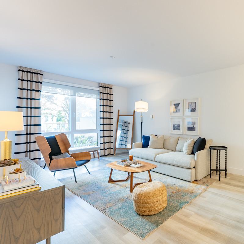 Modern apartment interior at The Triton featuring a sleek open kitchen with stainless steel appliances, light wood cabinetry, and a stylish navy blue sectional sofa beneath contemporary starburst pendant lighting.